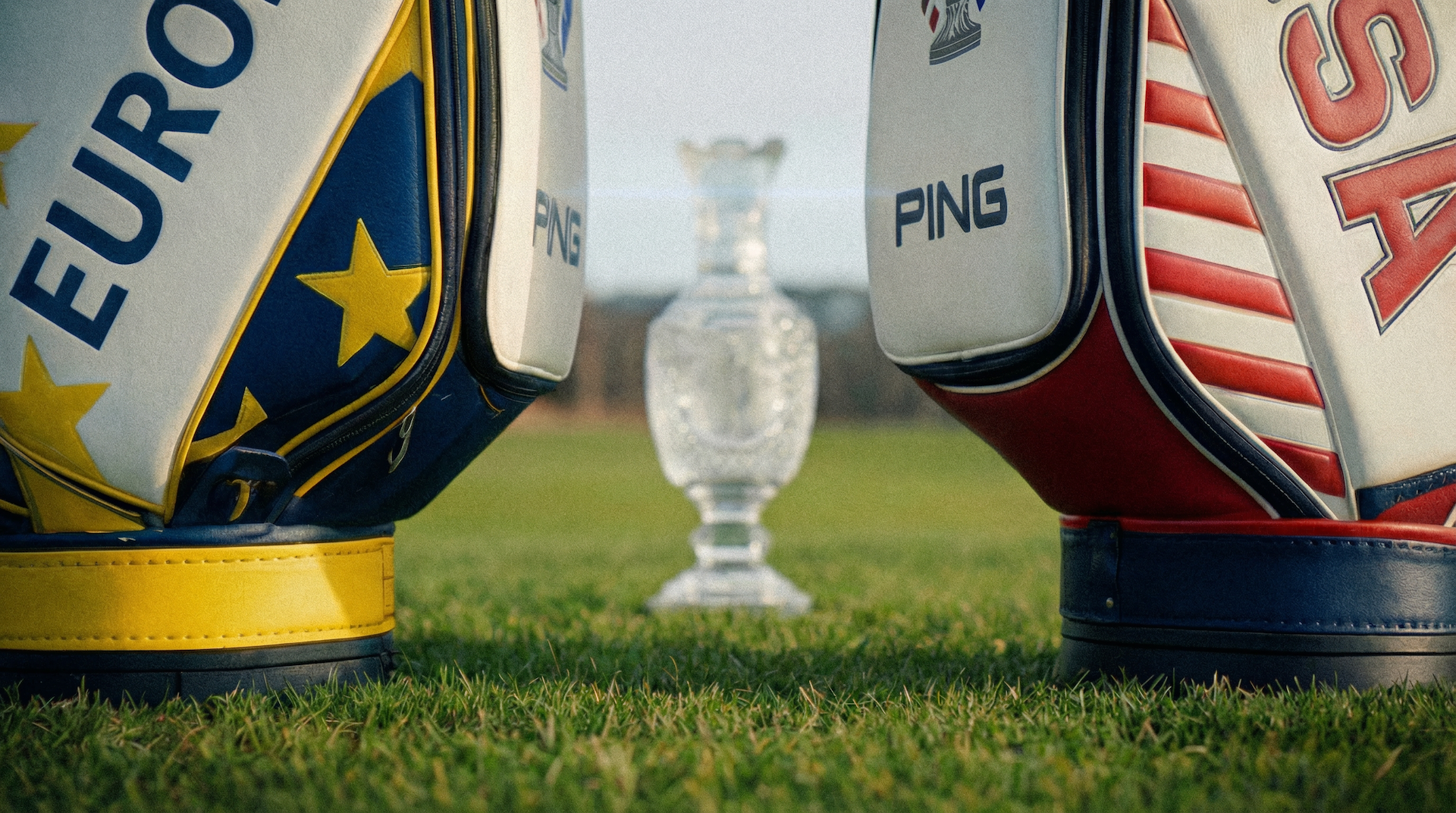 A view of two golf bags, one with a European Union flag design and the other with an American flag design, with a silver trophy in the background on a golf course.