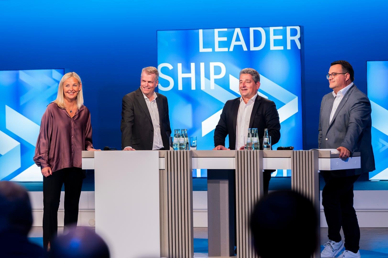 Four people standing behind podiums on a stage with a blue background and large text reading 'Leadership'.