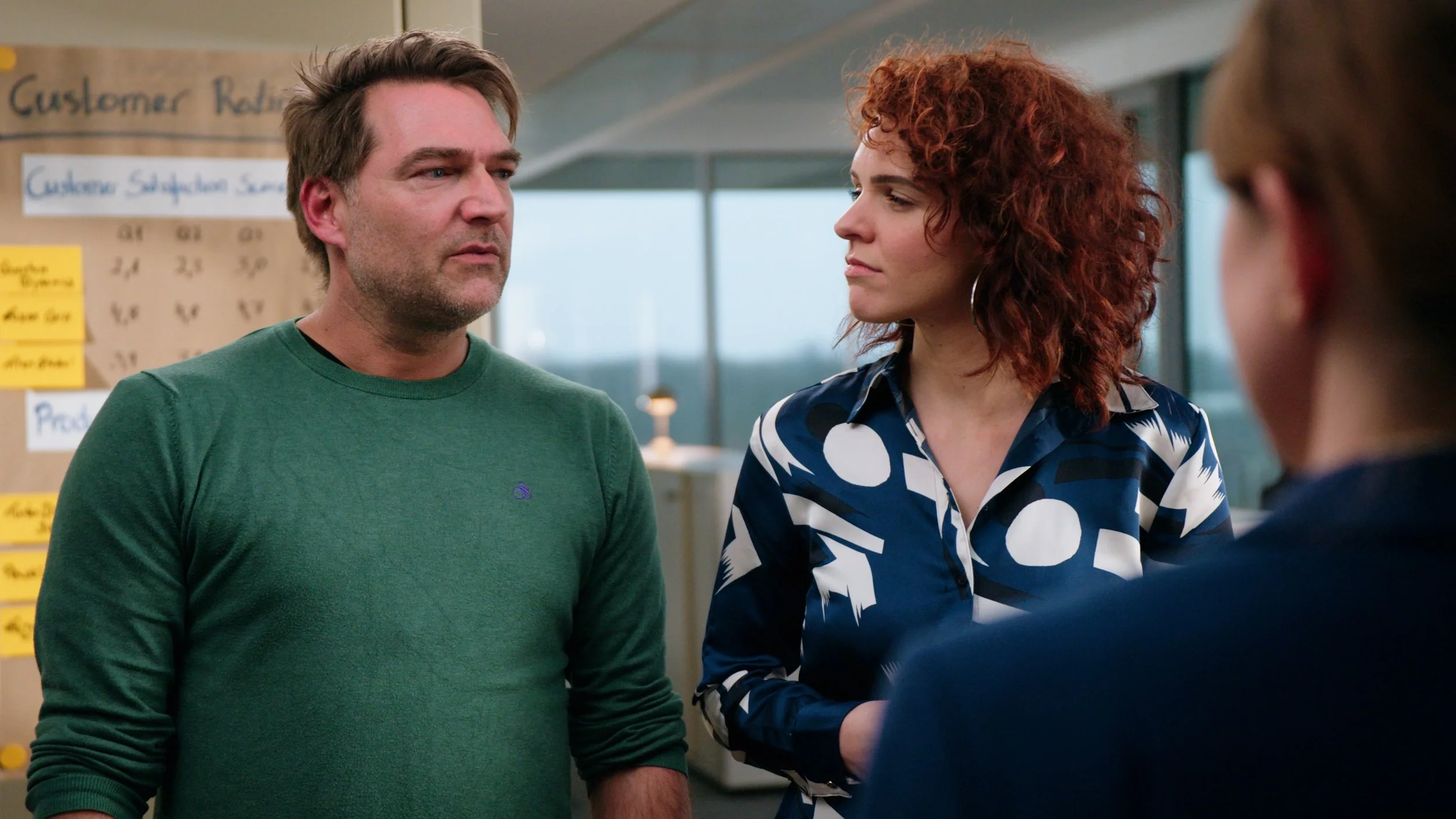 Three people having a serious conversation in an office setting, with aboard on the wall labeled 'Customer Relations' in the background.