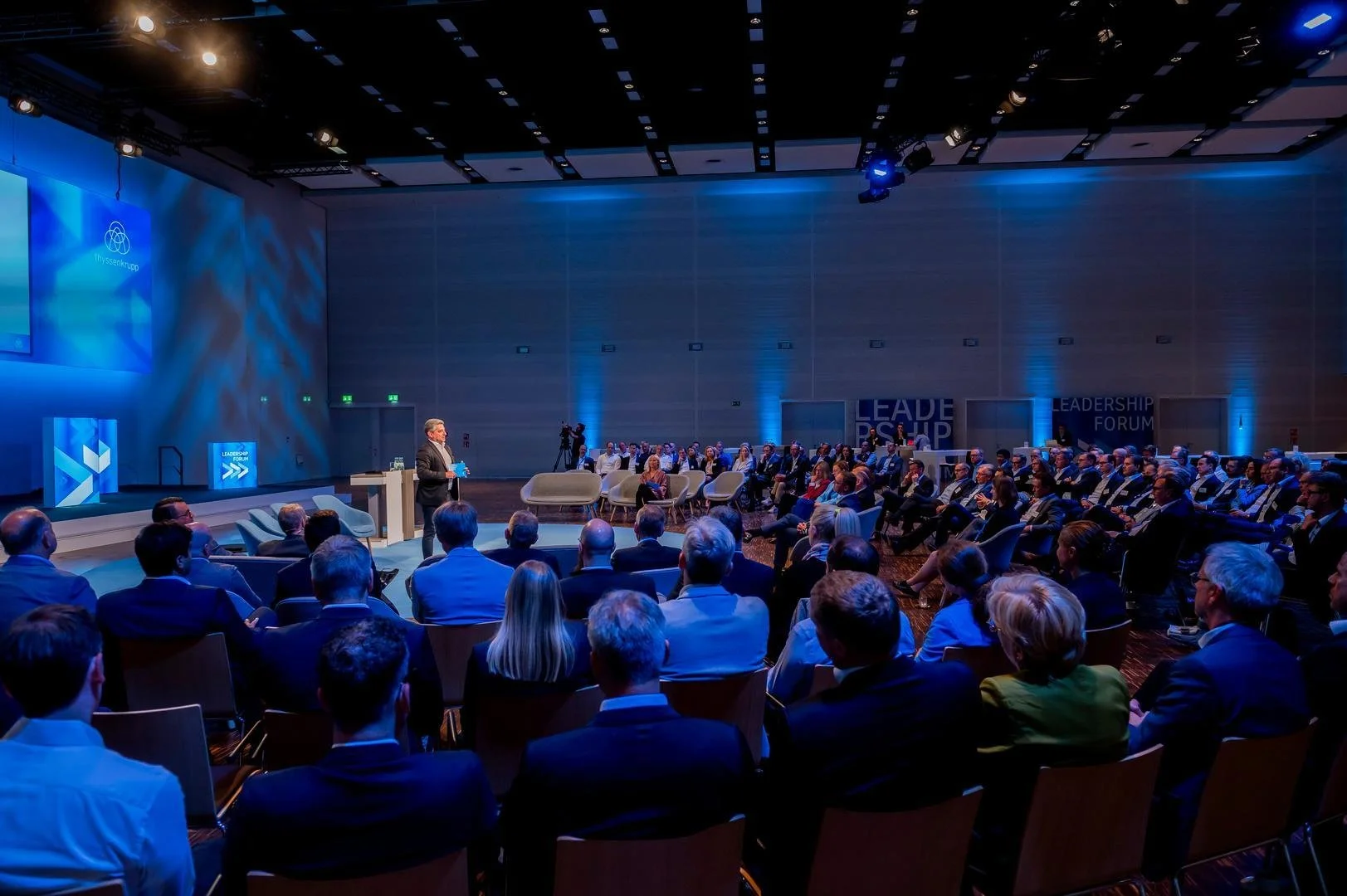 A conference or seminar with a speaker on stage addressing an audience seated in a large, dimly lit room with blue lighting and large screens displaying graphics.