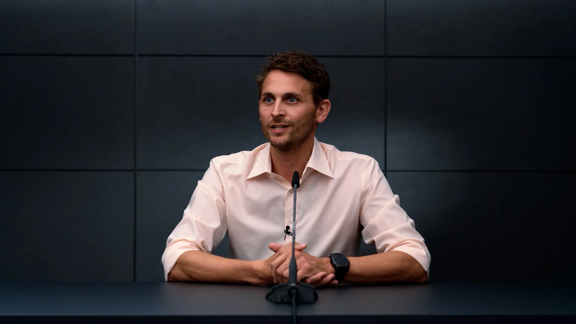 A man with dark hair and a beard sits at a table with a black microphone in front of him, wearing a light pink shirt and a black watch, against a dark tiled background.
