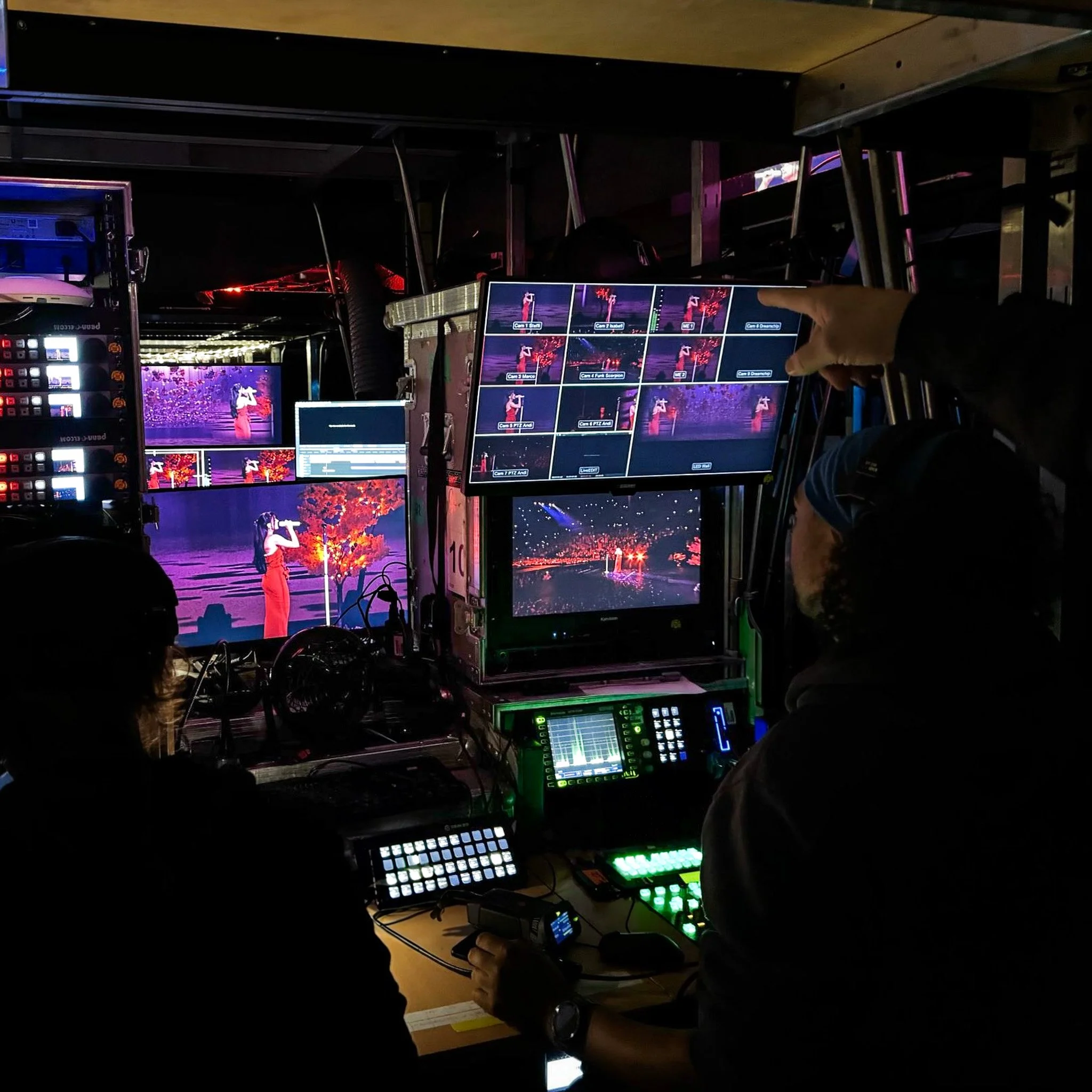 Behind-the-scenes view of a video production setup with multiple monitors, a control panel, and crew members filming a performer on stage with purple and red lighting.
