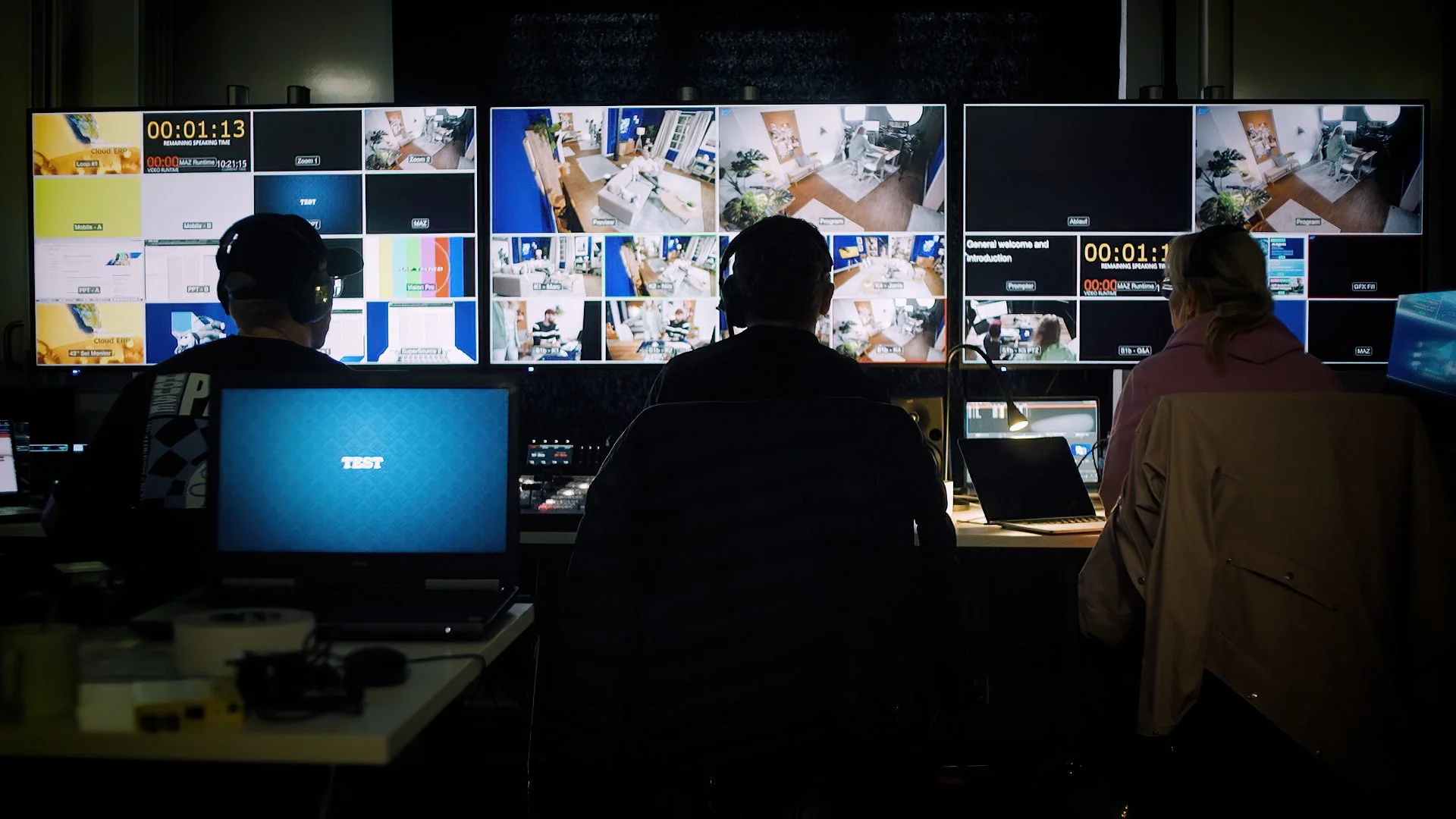 Three people sitting in front of multiple large screens displaying security camera footage and video channels in a control room.