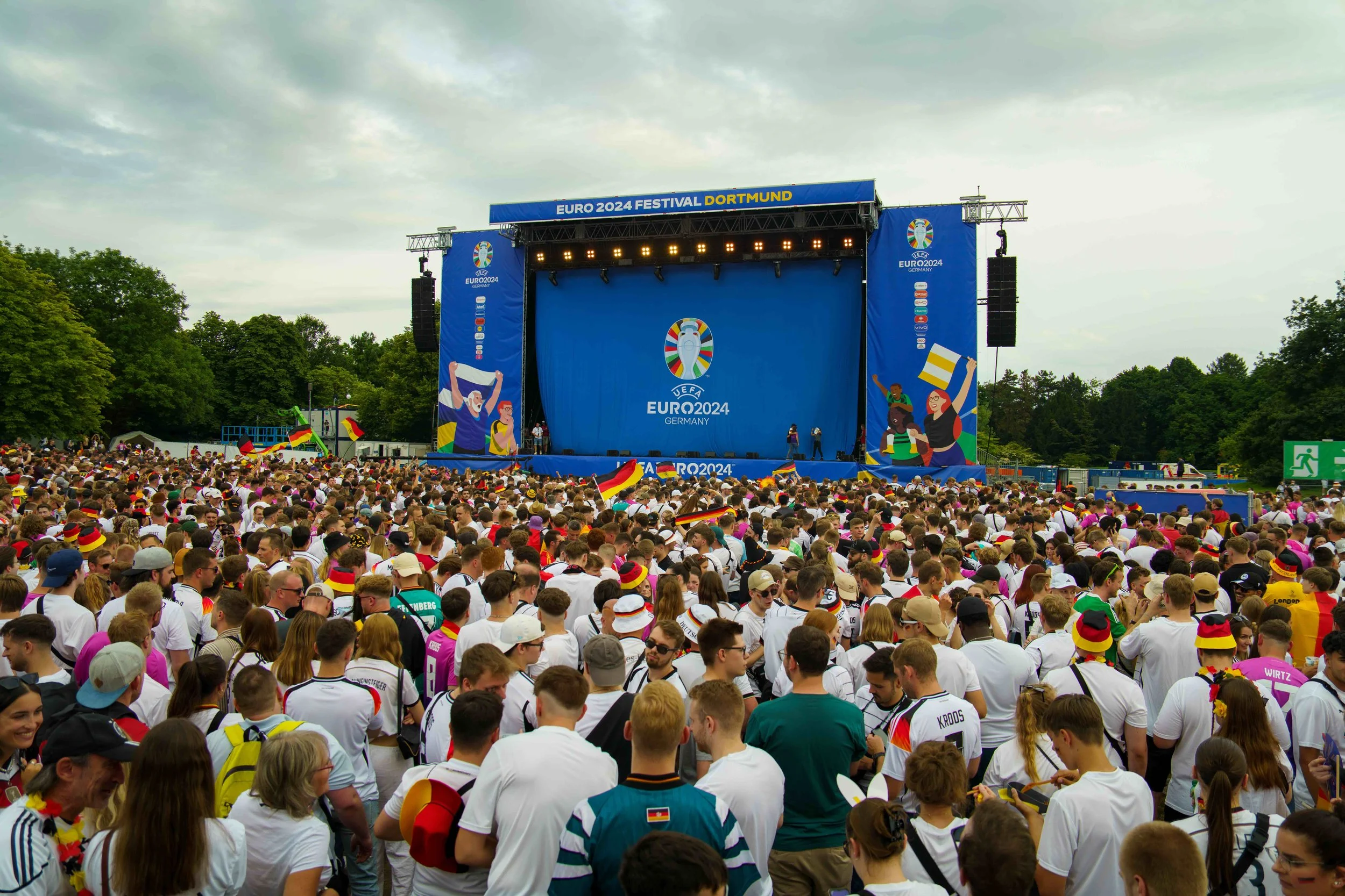 Crowd of people at an outdoor stage during the Euro 2024 Festival in Dortmund, Germany, with flags and sports apparel, and a large screen on stage with the Euro 2024 logo.