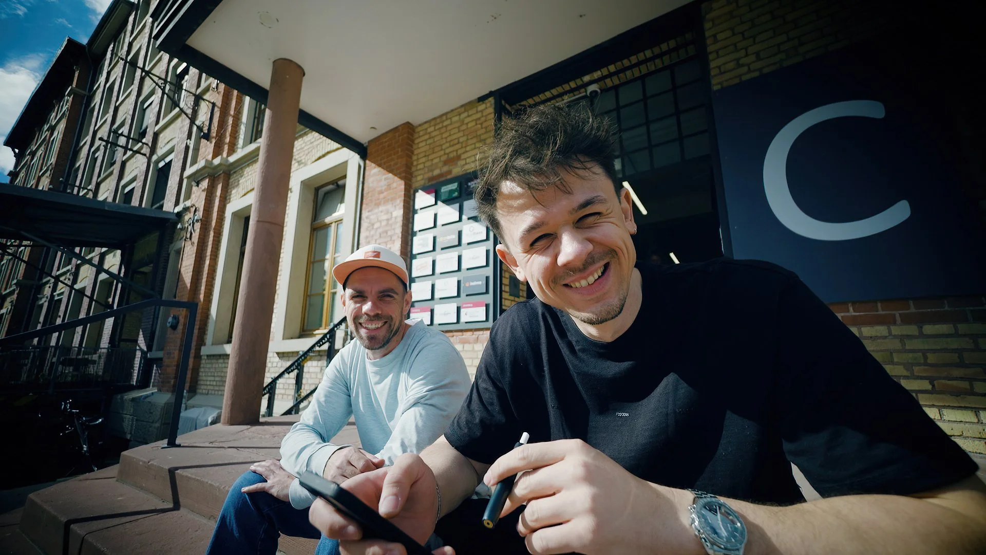 Two smiling men sitting on stairs outside a building, one holding a smartphone, with a large dark sign with a white letter C behind them.