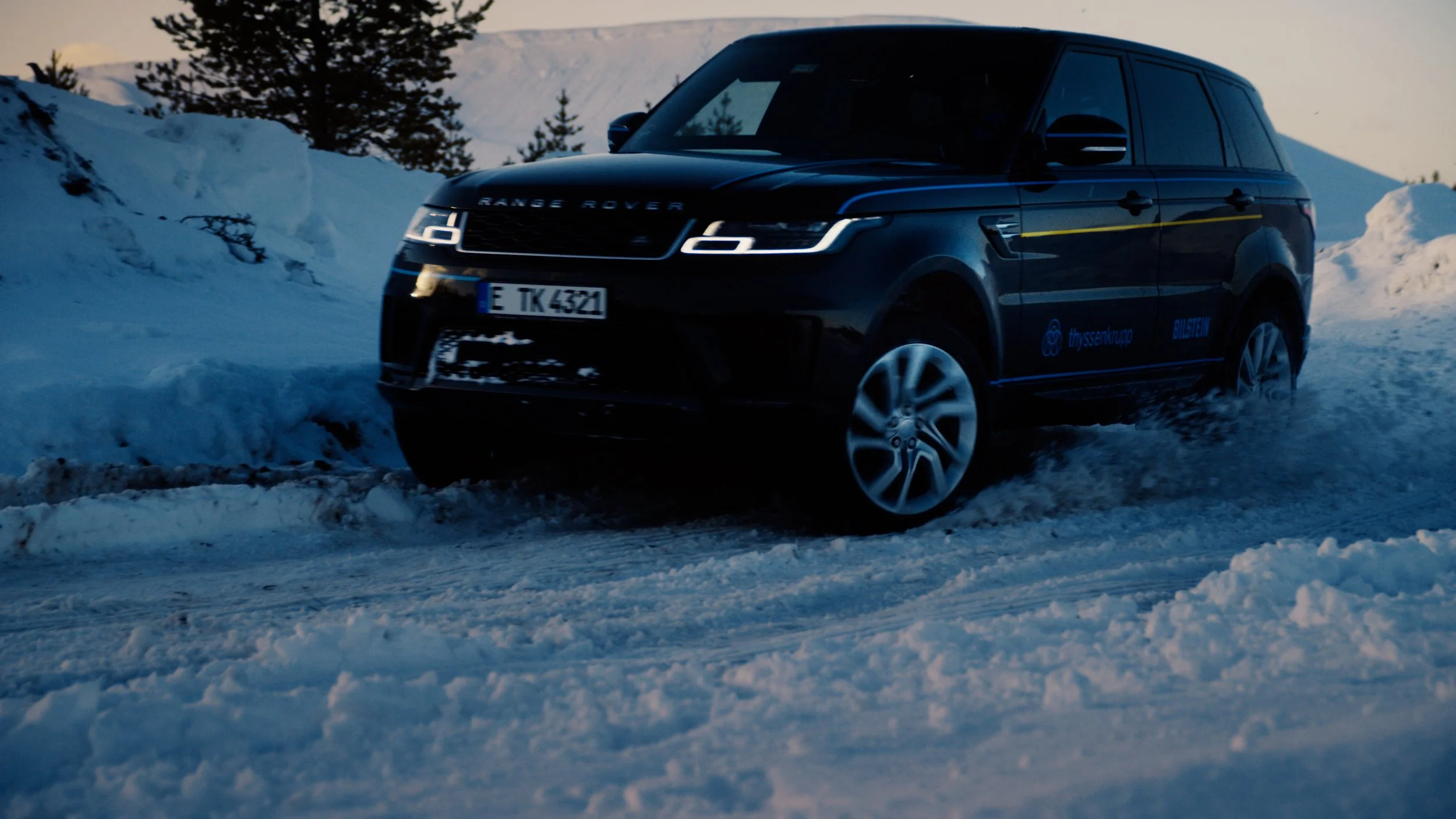 A black Range Rover driving through snow on a mountain road with snow-covered trees and mountains in the background.