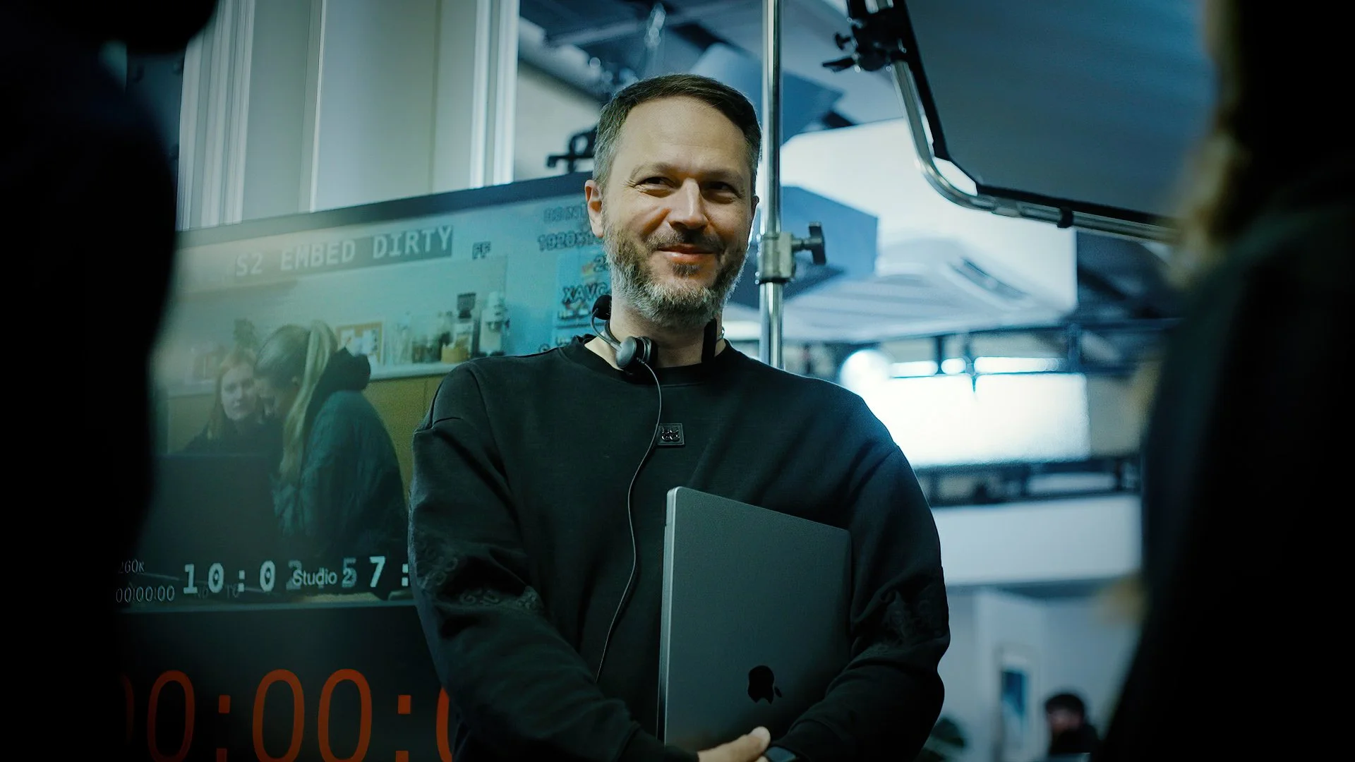 A man with a beard and short hair holding a laptop and wearing a headset, standing indoors with a screen and people in the background.
