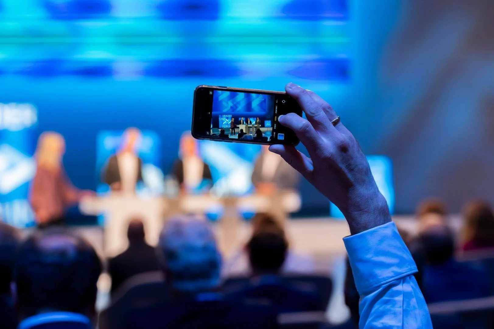 A person holding a smartphone in landscape mode, recording or taking a picture of a panel discussion on stage at a conference or forum with four speakers, in front of a large blue screen.