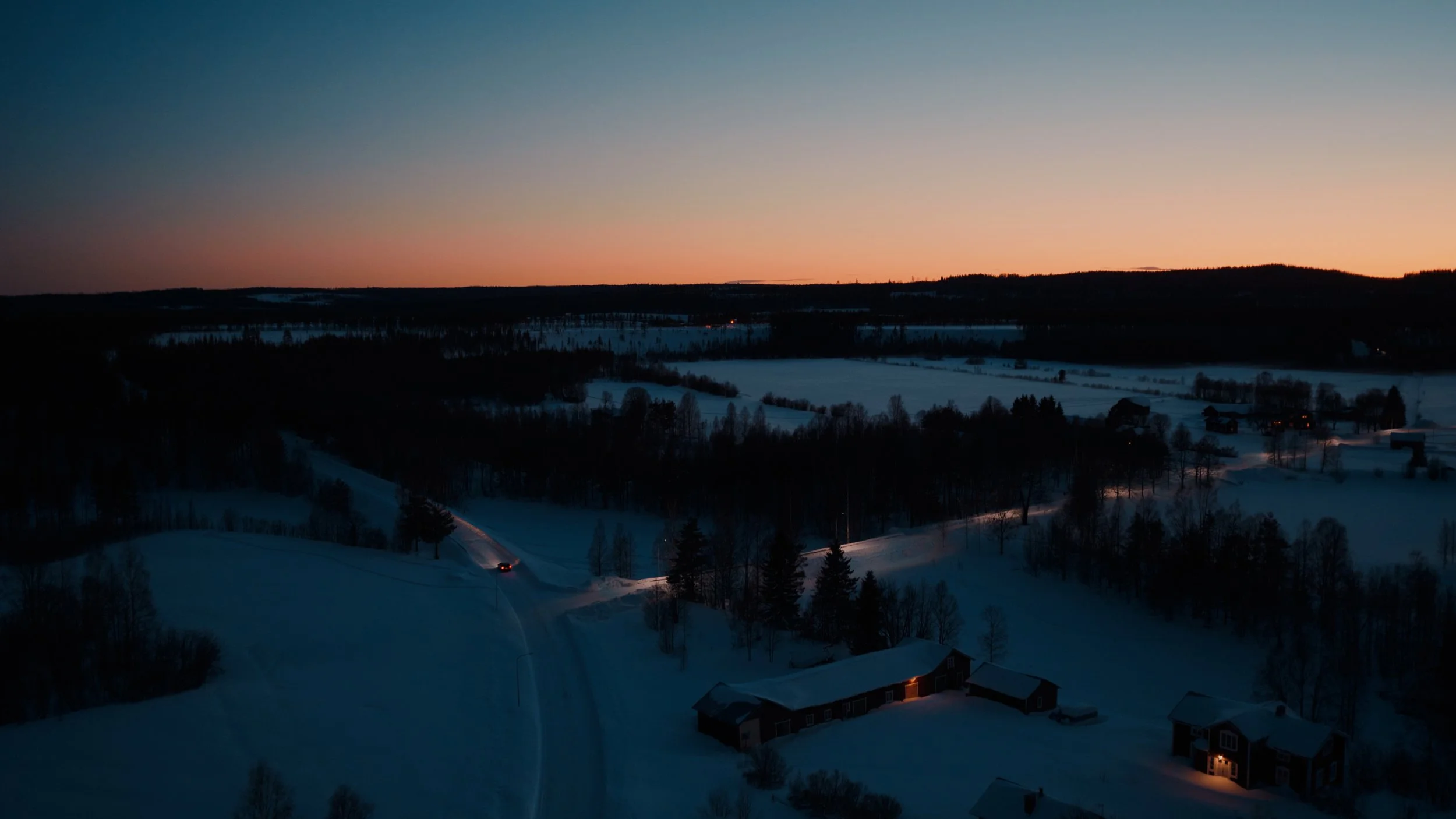 Snow-covered landscape at sunset with scattered houses and winding roads, hills in the distance, and a colorful sky transitioning from orange to dark blue.