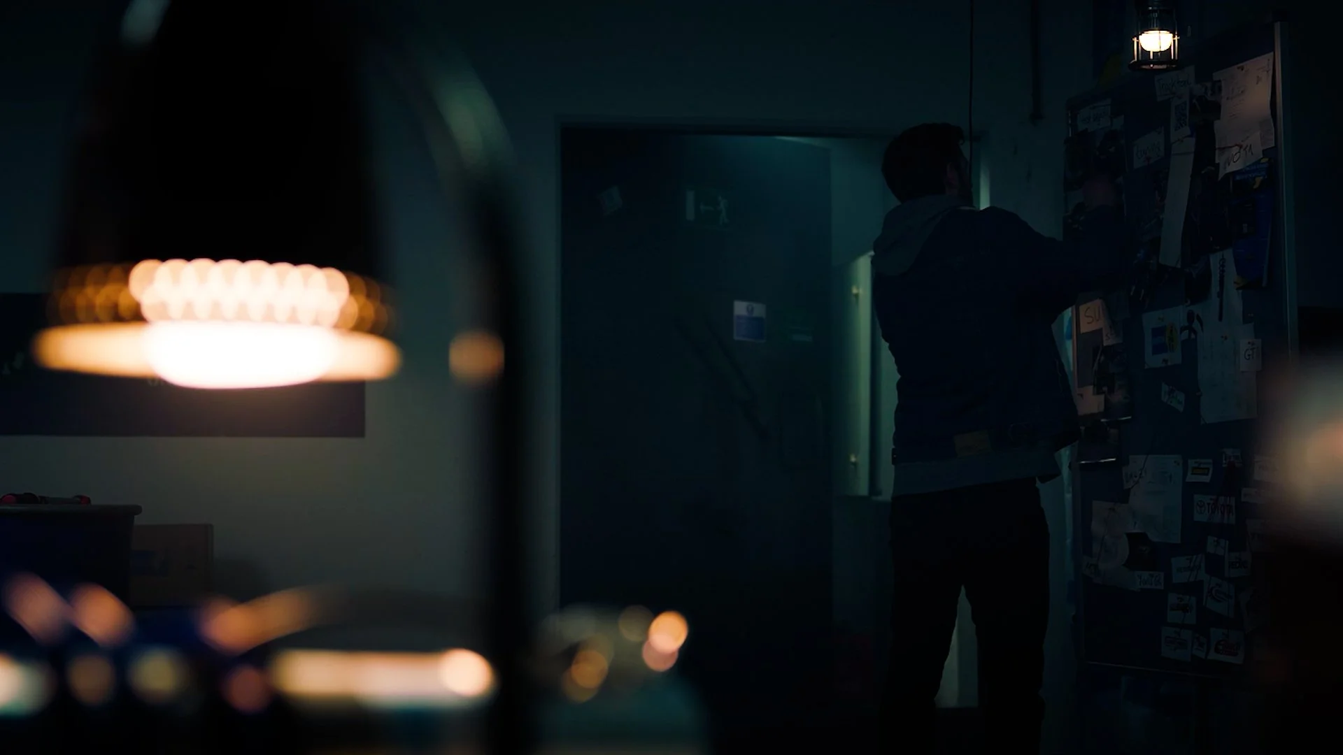 A person standing in front of a bulletin board covered with papers and stickers in a dimly lit room, with a hanging light and a blurred foreground of lights.