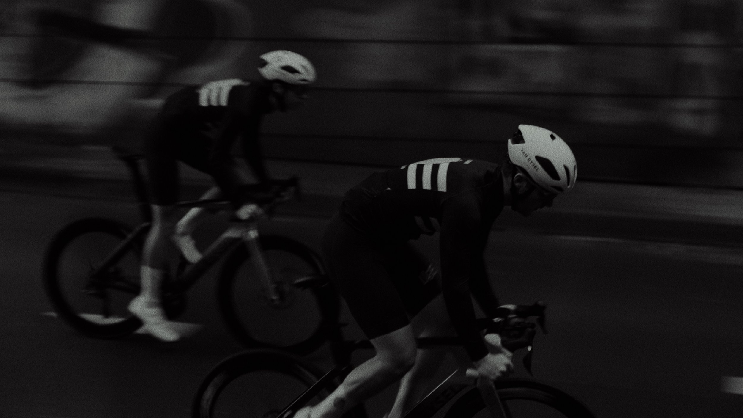 Two cyclists wearing helmets riding on a road at night in black and white. The front cyclist is leaning forward, while the one behind is slightly elevated.