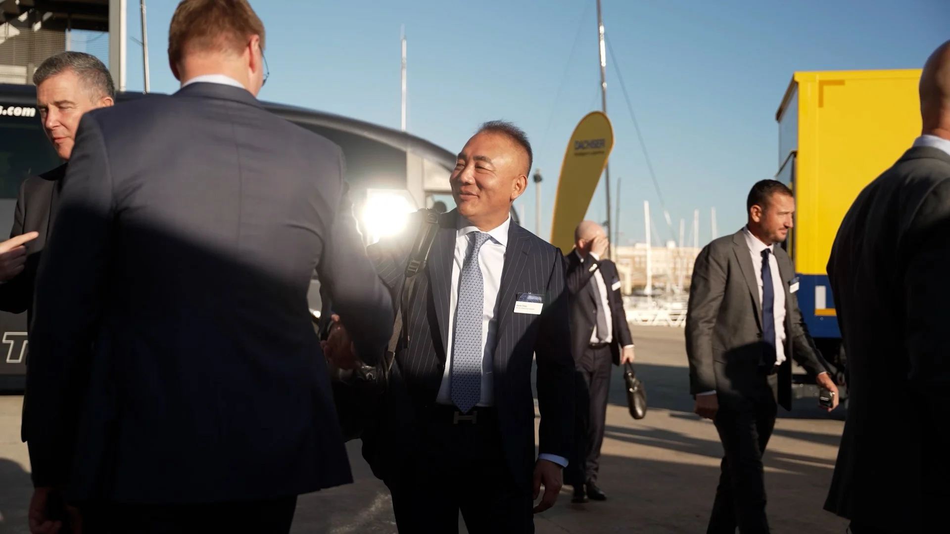 A group of people in business attire at an outdoor airport or marina, conversing and walking near a black vehicle, with boats and clear sky in the background.