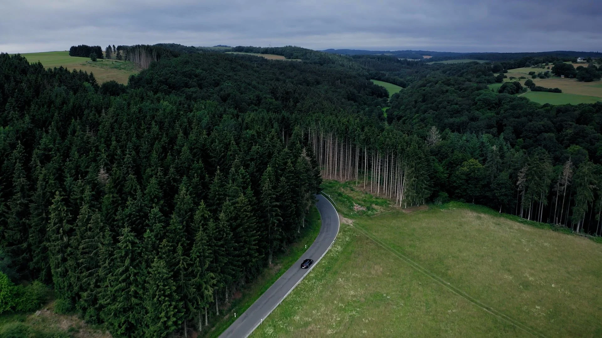An aerial view of a winding mountain road passing through a dense forest with a solitary dark-colored car driving on it, surrounded by green trees and open grassy fields under a cloudy sky.