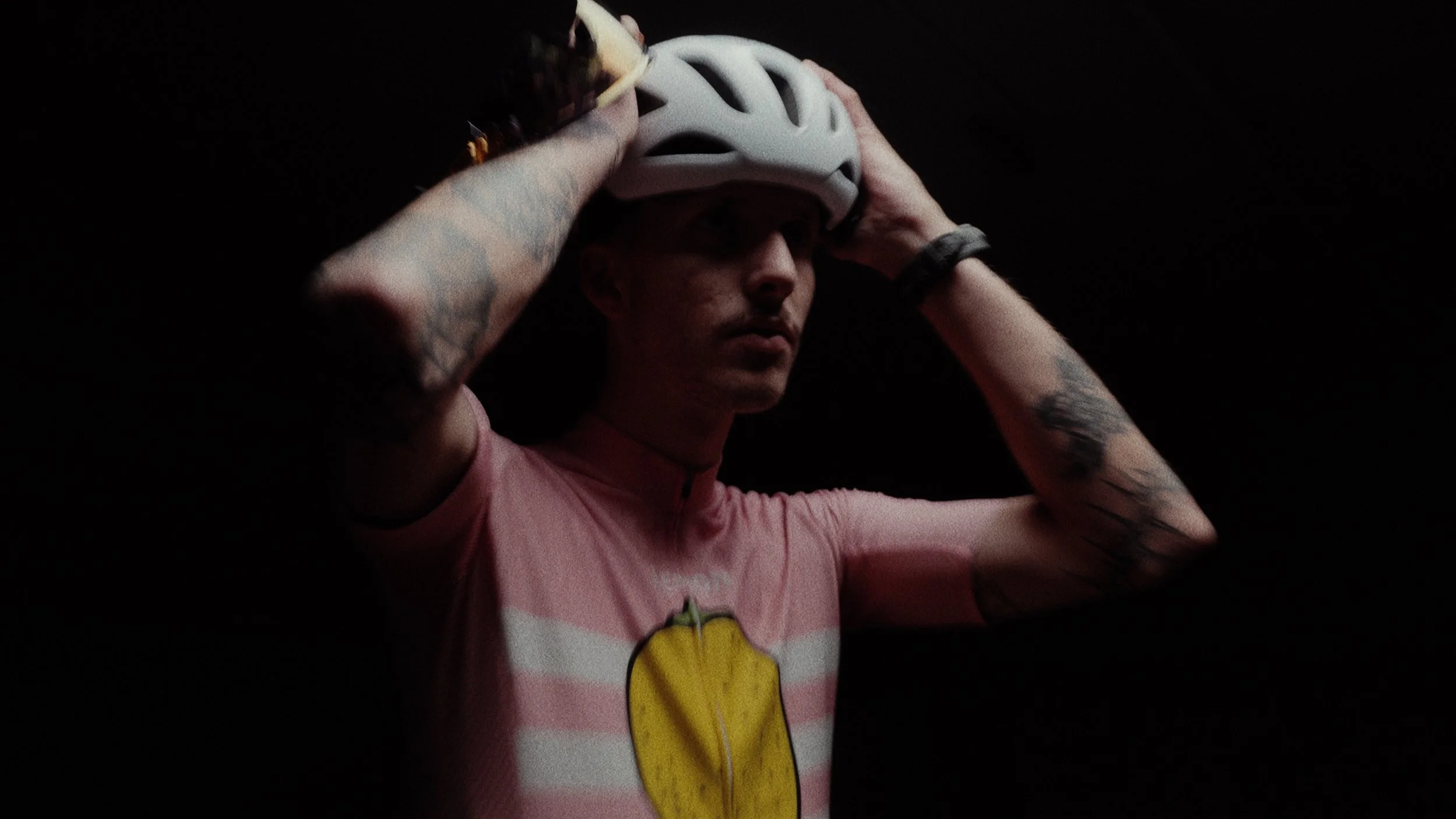 A person with tattoos on their arms wearing a pink and white striped shirt with a lemon graphic, holding a white bike helmet on their head against a dark background.