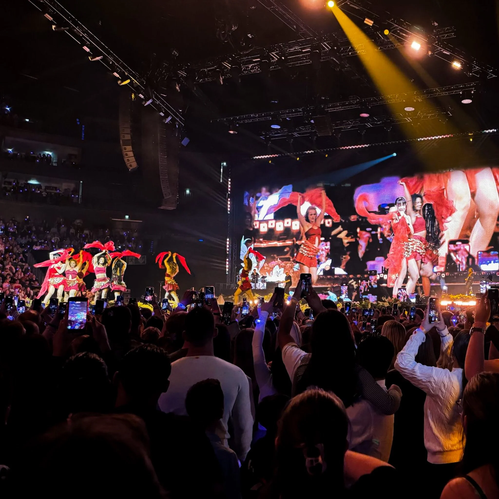 A concert with a large crowd watching a stage performance featuring dancers in red costumes and large digital screens showing the performance.