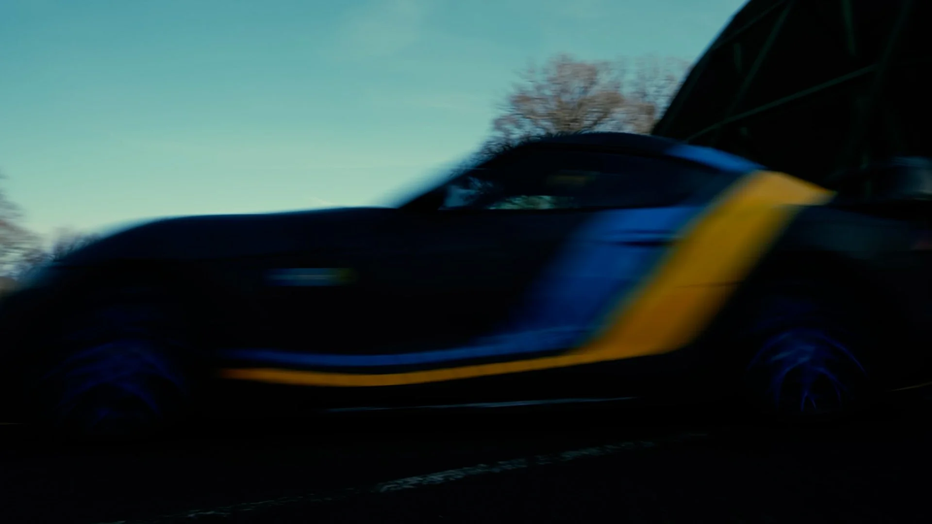 A close-up of a black sports car with yellow accents, speeding on a road during daytime. Trees are visible in the background under a clear blue sky.