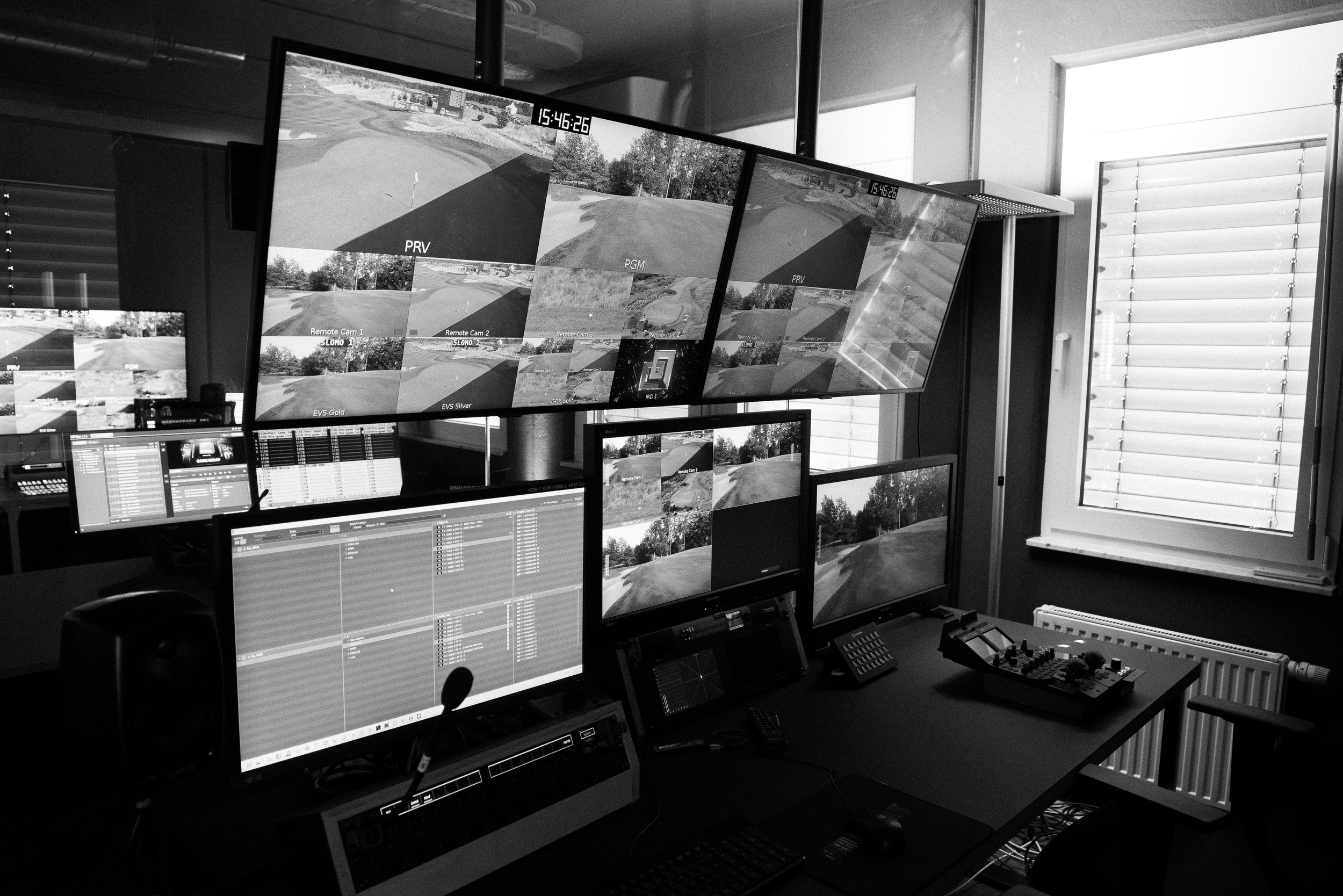 Multiple monitors displaying surveillance footage of a golf course, with a control desk and various electronic equipment in a room with a window and blinds.