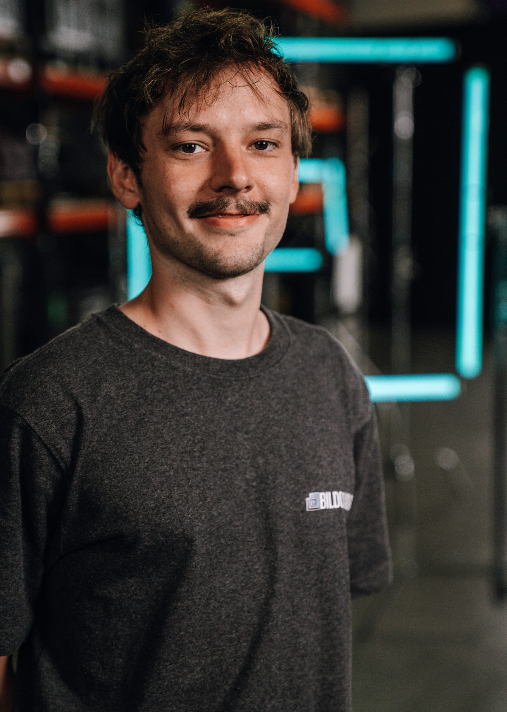 A young man with a mustache and brown hair, smiling, wearing a dark gray T-shirt, standing in front of a backdrop with neon lights.