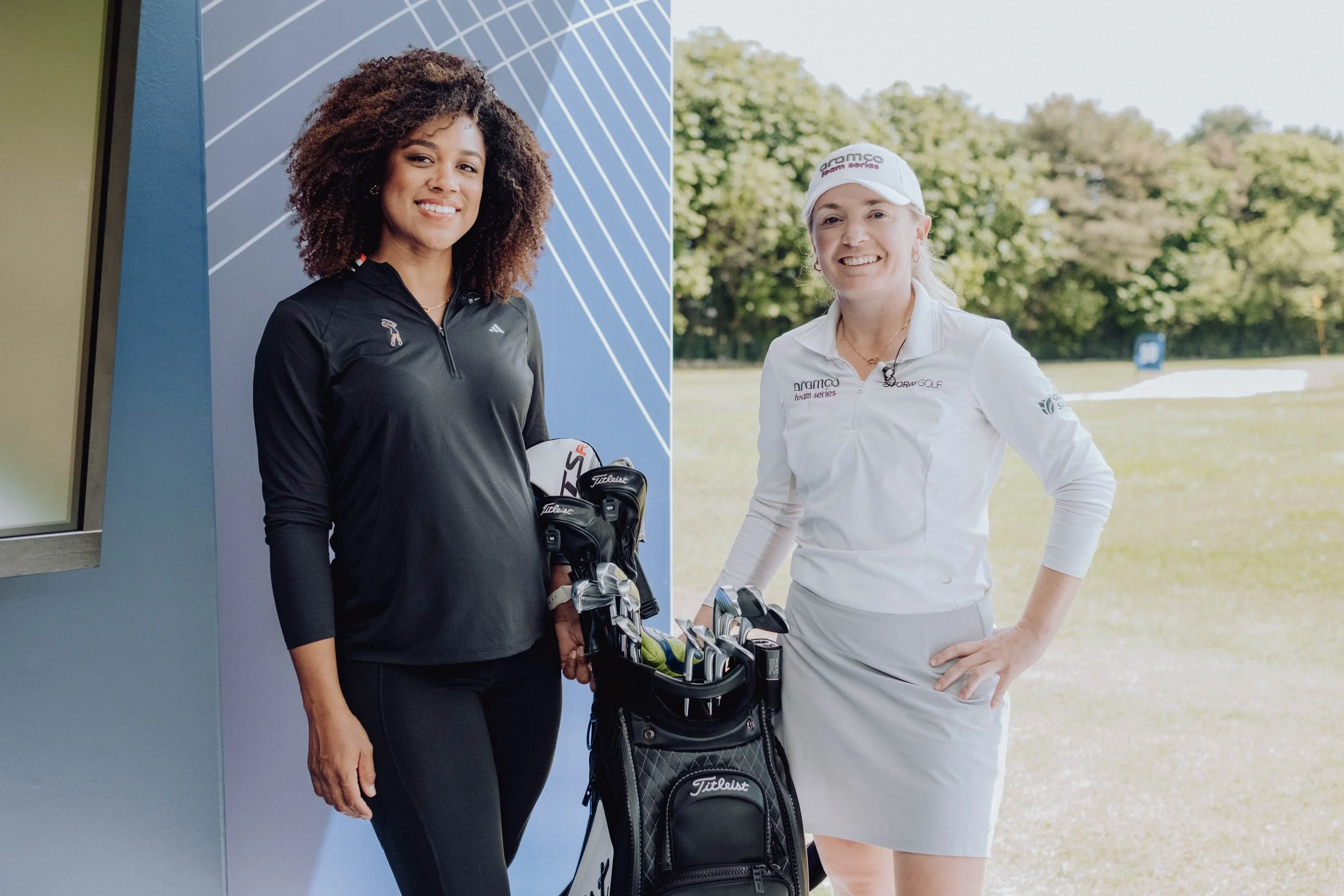 Two women on a golf course, one dressed in black holding golf clubs, and the other in white with golf clubs in her bag, smiling at the camera.