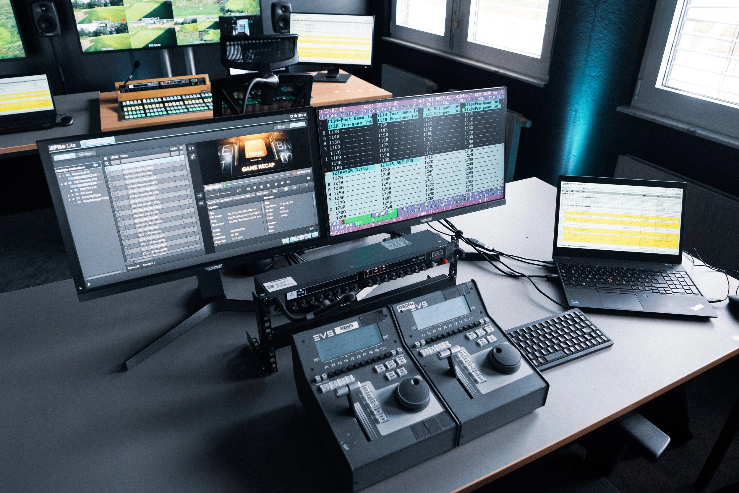 A video production control room with multiple monitors displaying editing and scheduling software, audio mixing hardware, and a laptop with spreadsheets, all on a long desk.