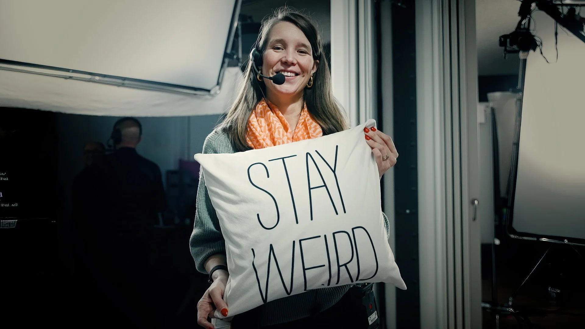 A woman with long brown hair, wearing a gray sweater, orange patterned scarf, and headset, smiling and holding a white pillow with the words 'STAY WEIRD' written on it.