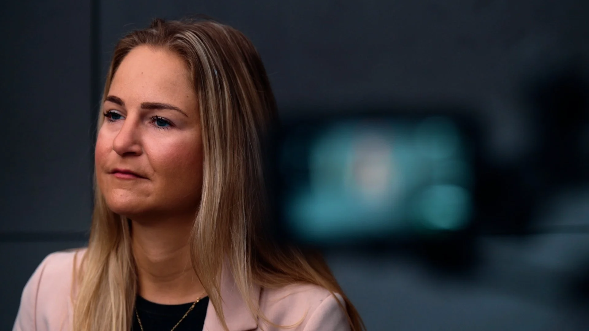 A woman with long blonde hair and blue eyes looking off to the side, standing against a dark background.