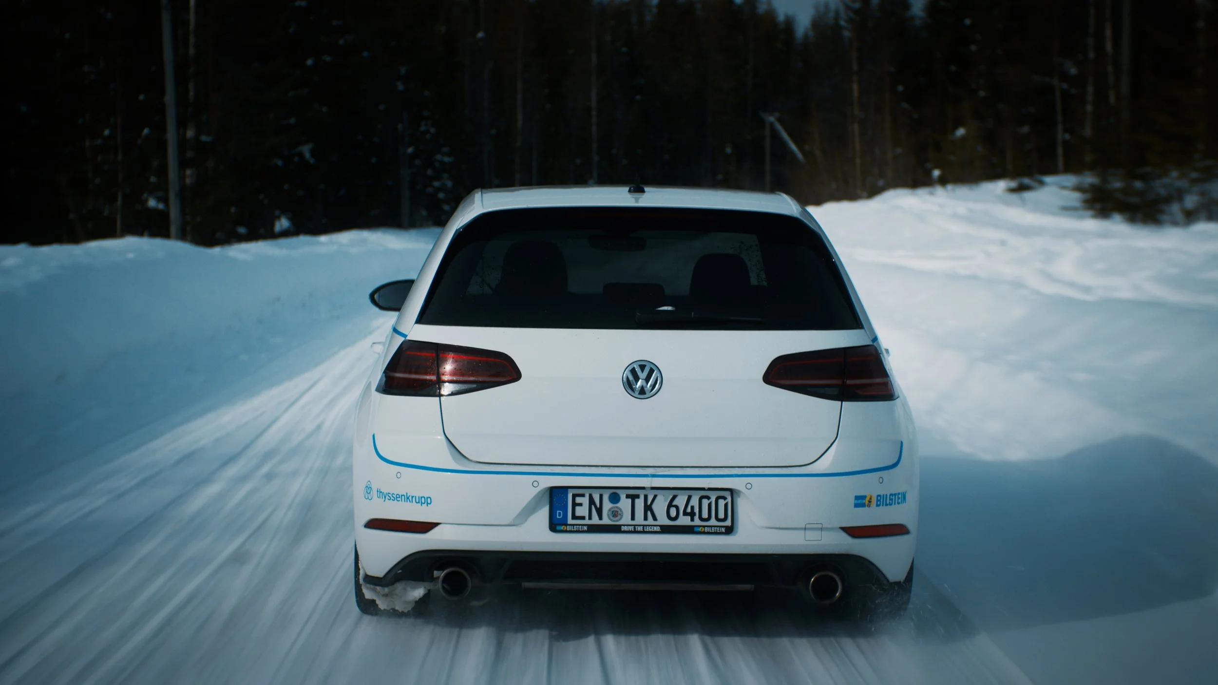 White Volkswagen car driving on a snowy road, surrounded by snowbanks and dark forest in the background.