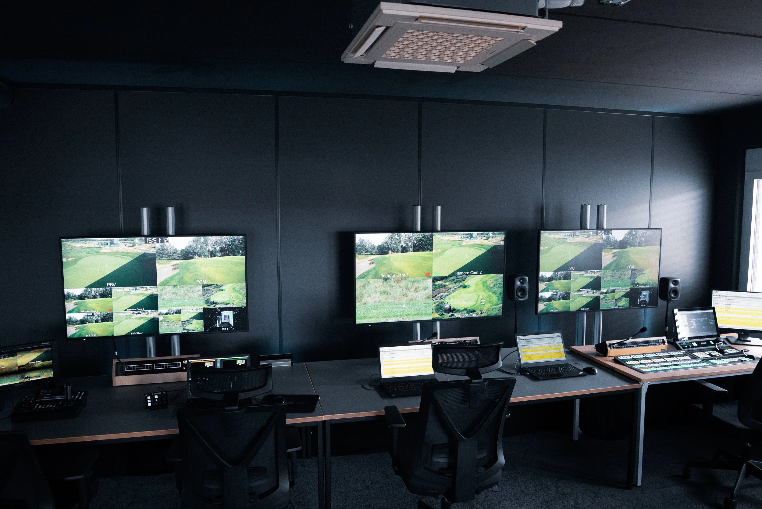 Control room with three large monitors displaying golf course footage, desks with laptops, audio equipment, and chairs, in a dark room.