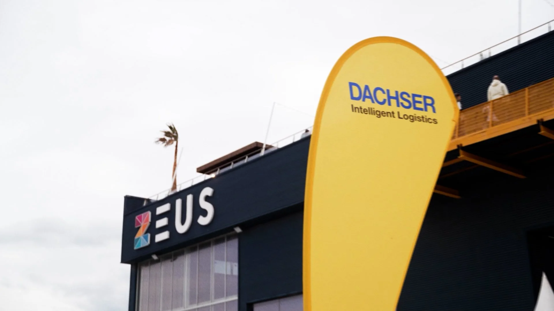 Yellow teardrop-shaped sign with blue text that reads 'DACHSER Intelligent Logistics' in front of a modern black building with a ZEUS sign on it. There are a few people on an elevated balcony and a palm tree in the background, with a cloudy sky overh