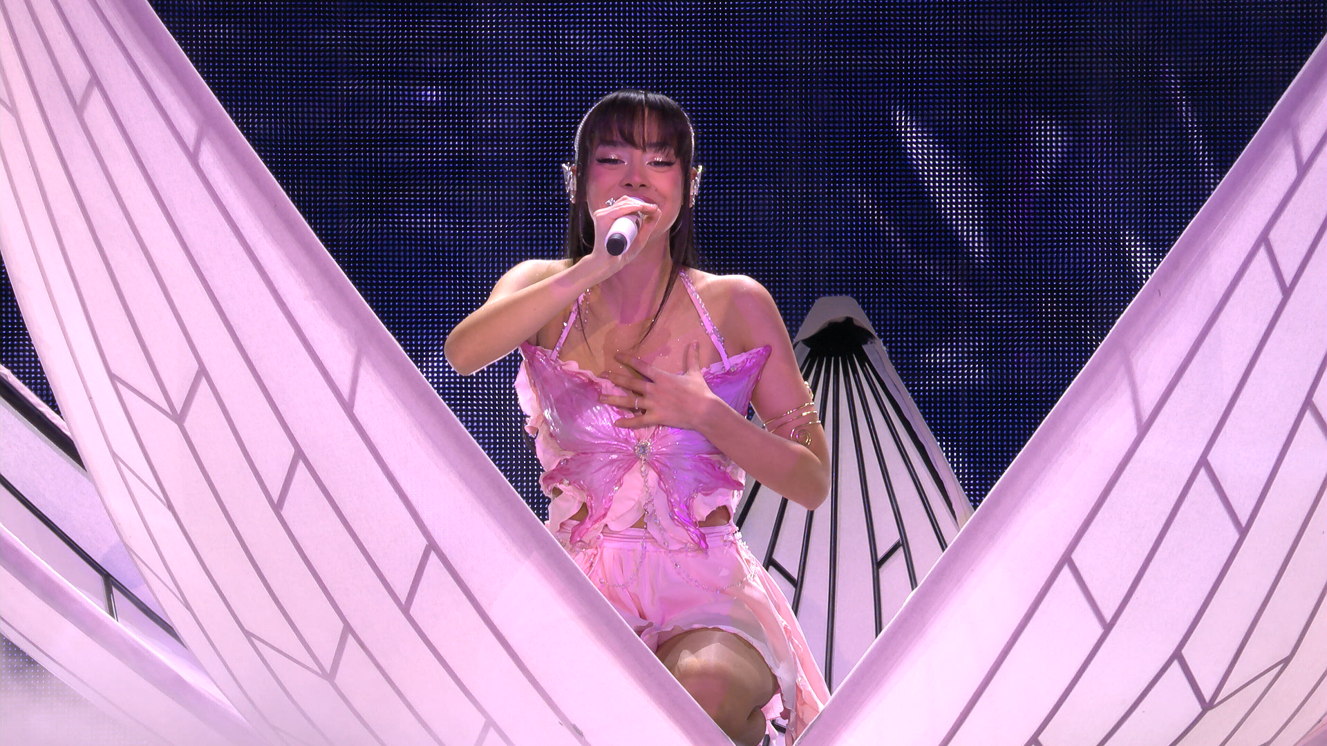 A young woman performing on stage, dressed in a pink butterfly-themed outfit, singing into a microphone with one hand on her chest and eyes closed.