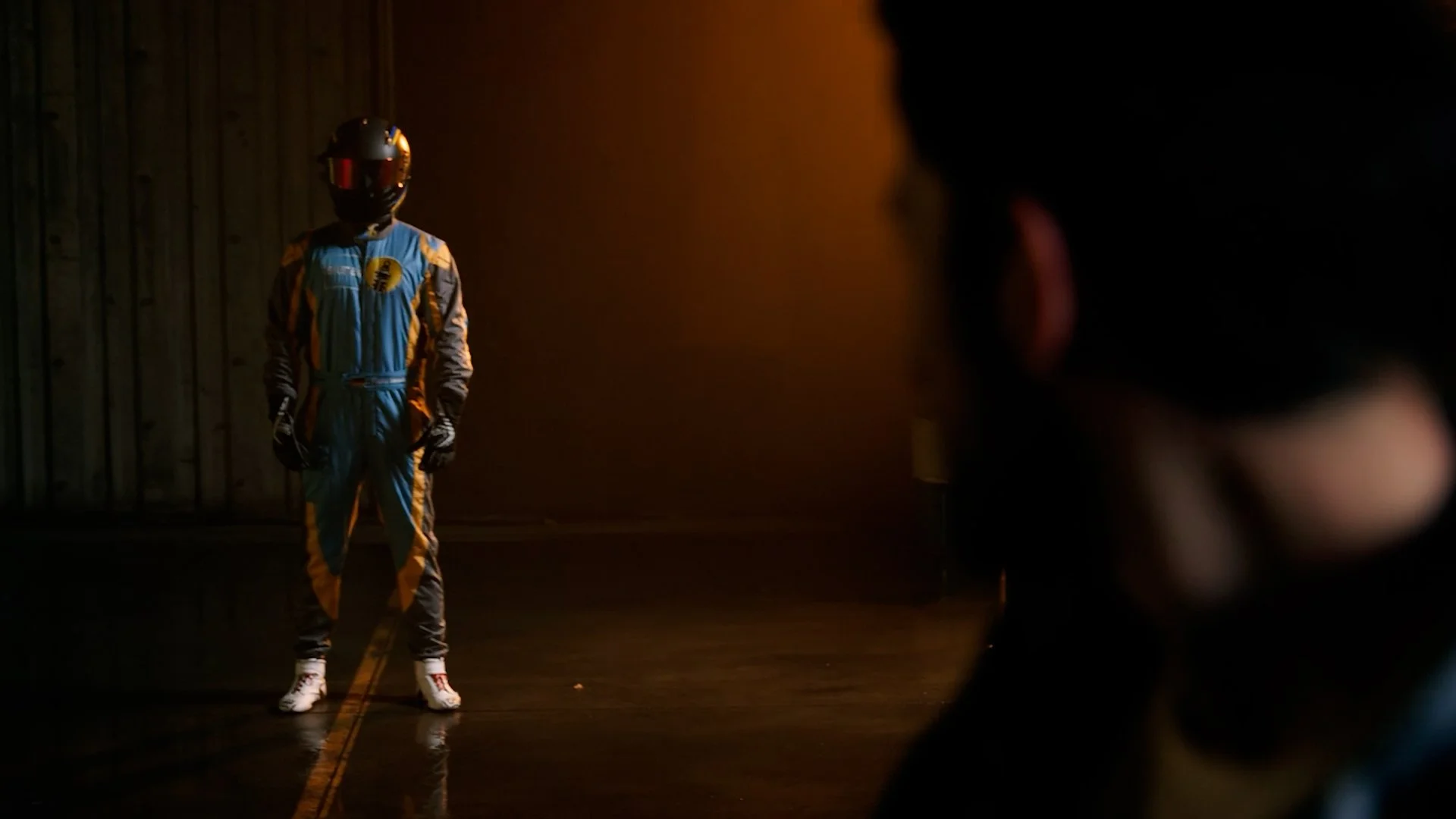 A race car driver in a racing suit and helmet standing on a dark, reflective surface in front of a wooden wall, with a blurred person or object in the foreground on the right side.