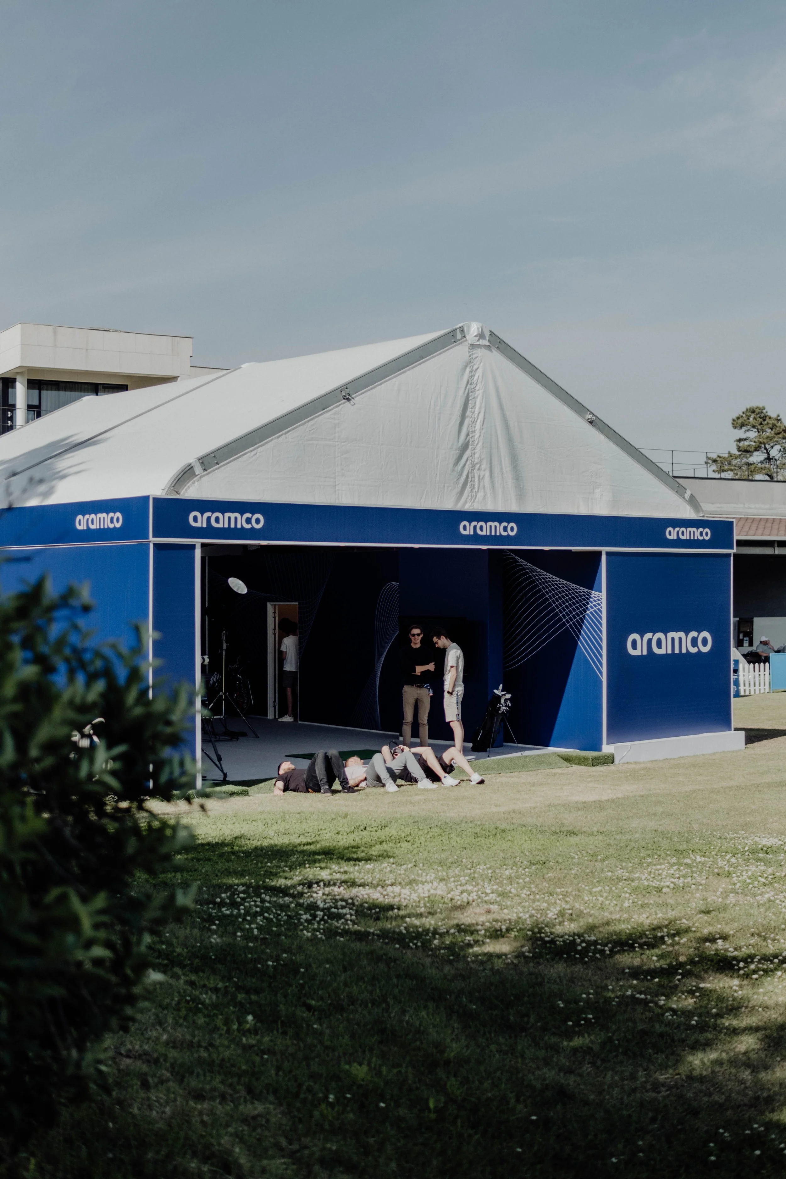 A large white event tent with a blue band featuring the 'Aramco' logo, set up on a grassy outdoor area with a partly cloudy sky. Several people are standing and lying on the grass near the tent, which has a stage or entrance area.