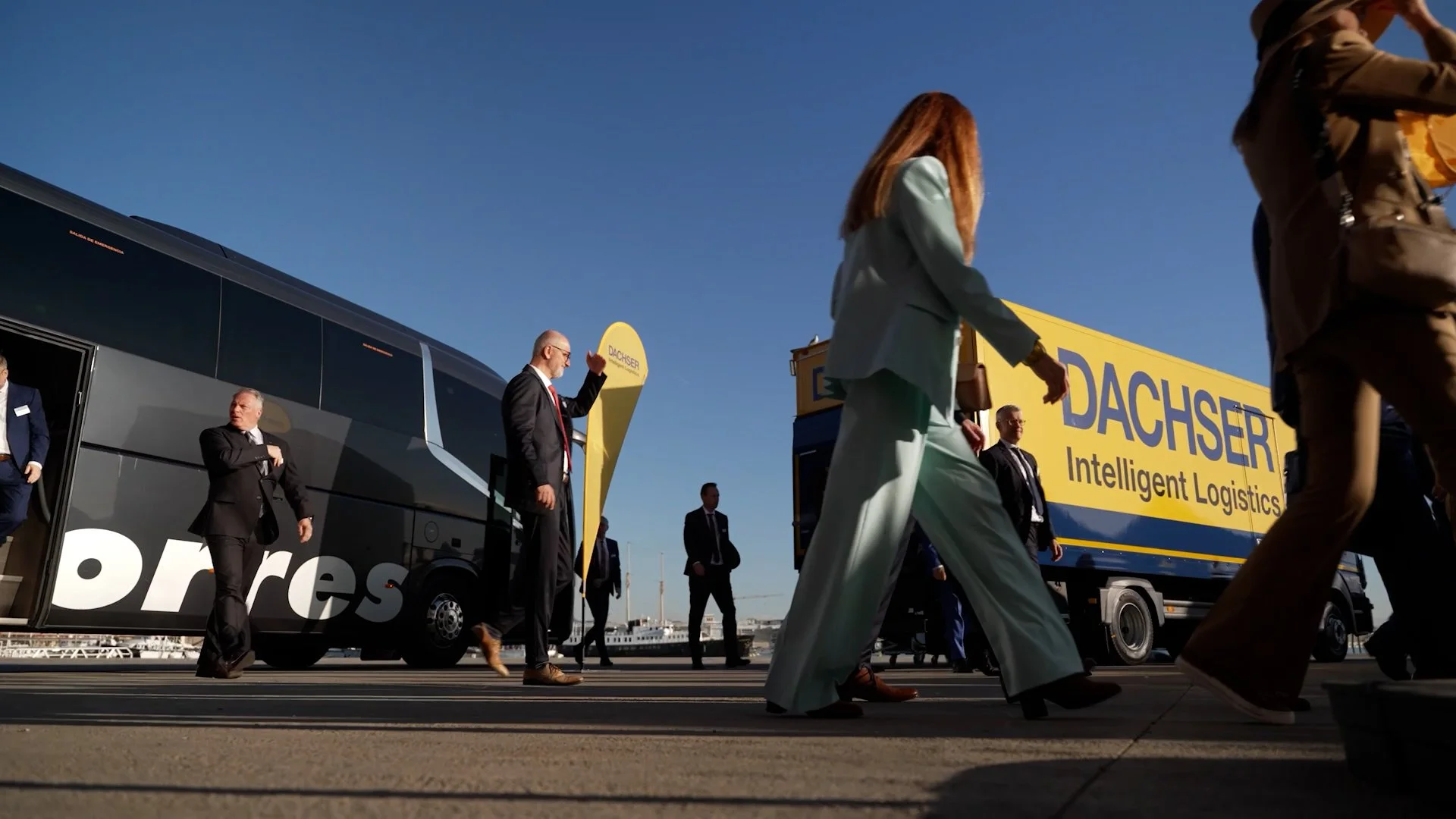 People walking outside near a large black bus and an orange, yellow, and blue truck with the words "DACHSER Intelligent Logistics" on it. The sky is clear and blue.