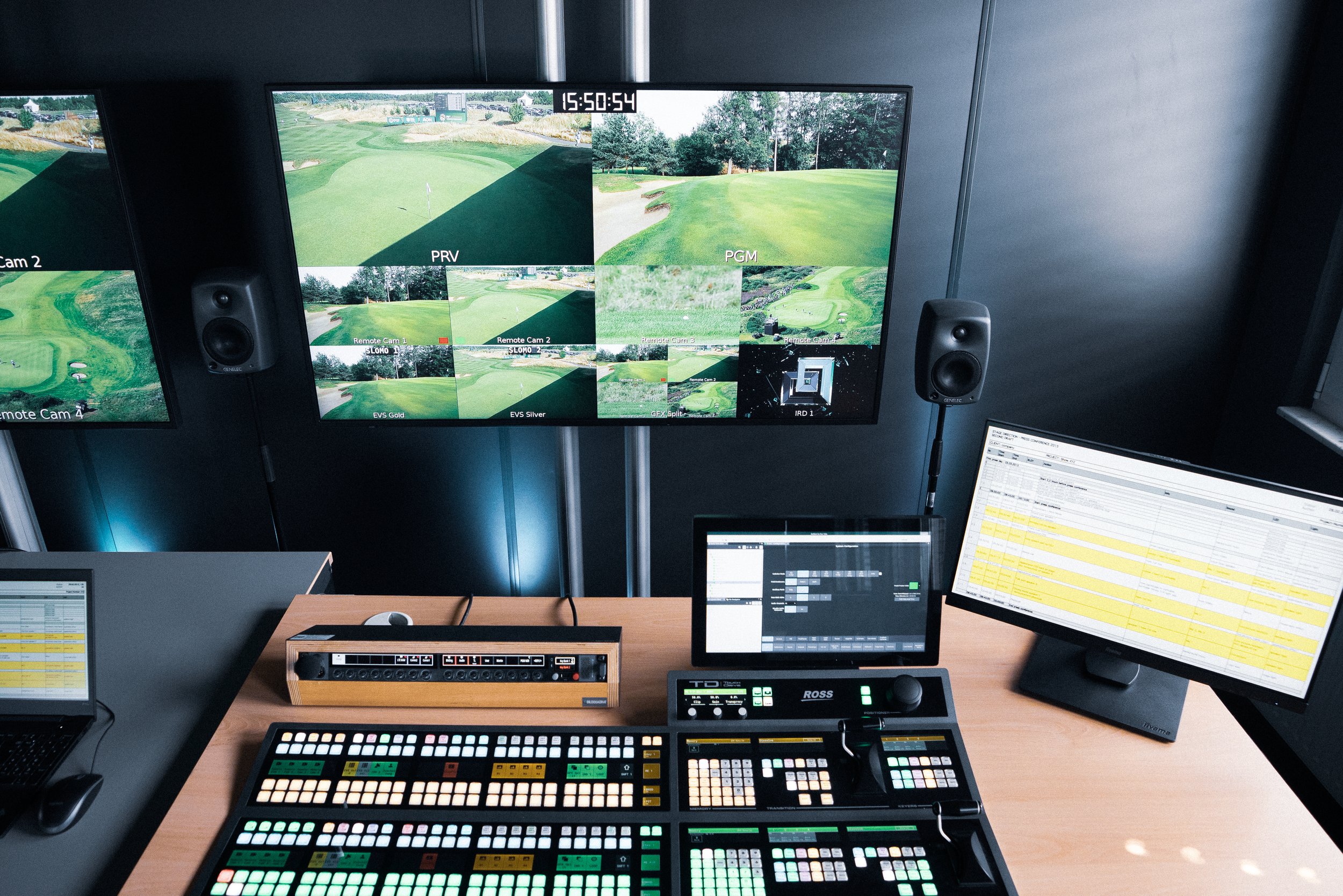 A control room with multiple screens showing live golf course feeds, a mixing console, and computer monitors displaying a scheduling or editing interface.