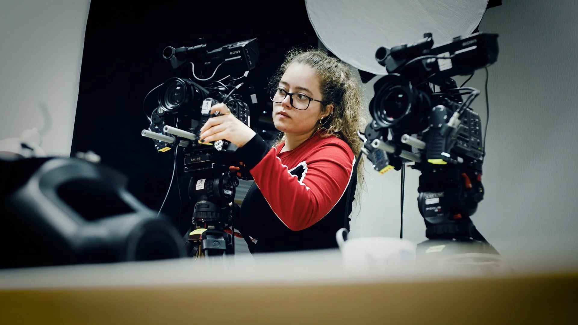A woman with glasses and curly hair operating a professional video camera in a recording studio.