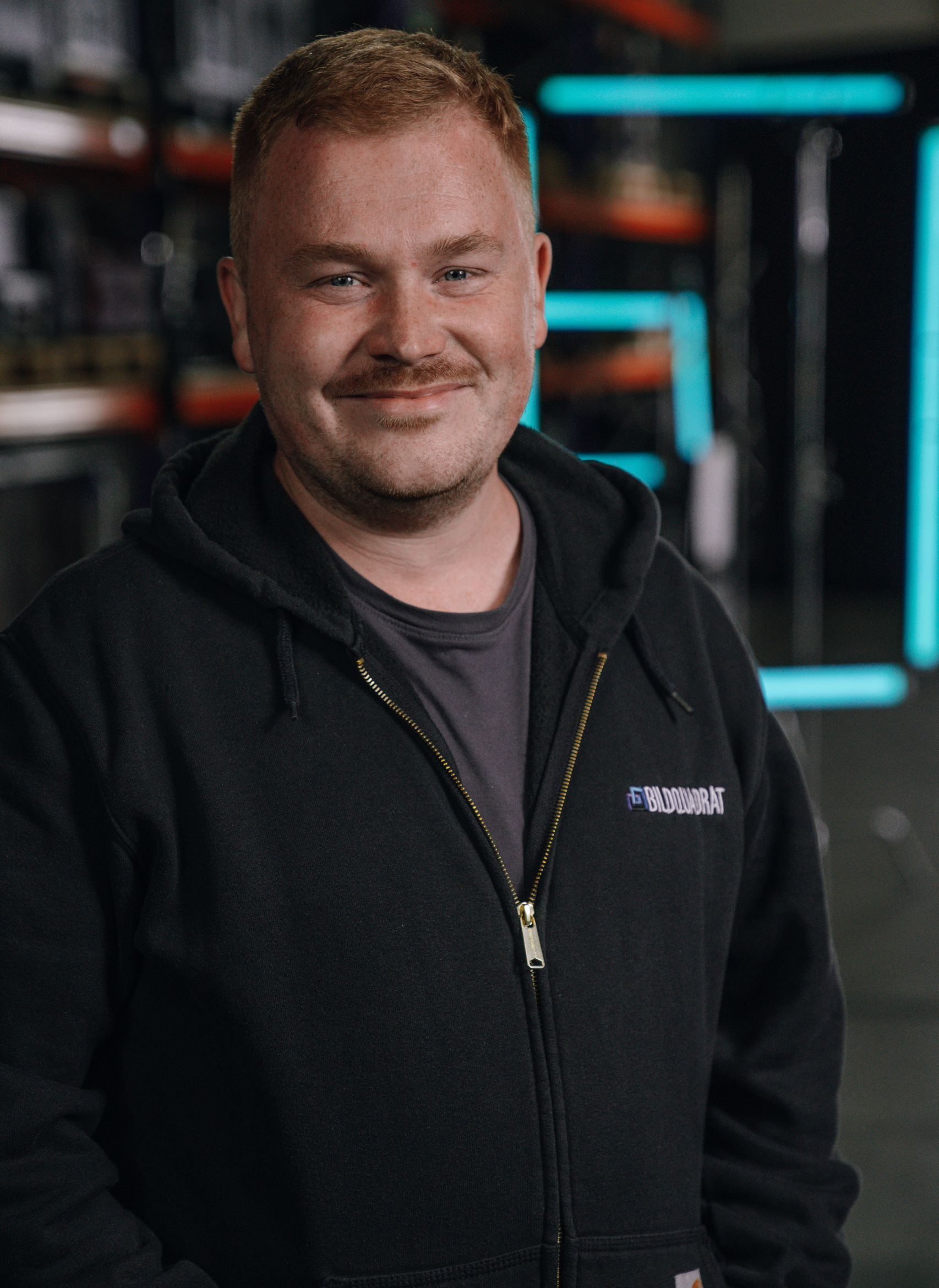 A smiling man with short red hair and a beard, wearing a black zip-up hoodie with a logo, standing in a warehouse or workshop environment with shelves and neon lights in the background.