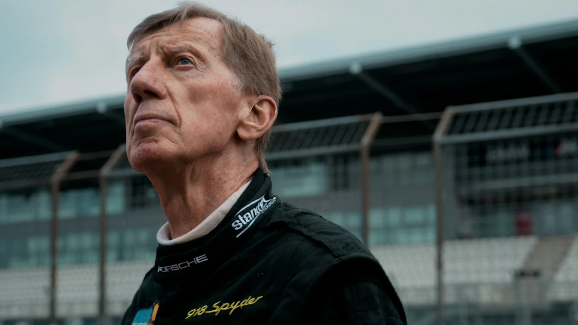 A man in a racing suit with the Porsche logo, standing near a race track, looking upwards with a serious expression.