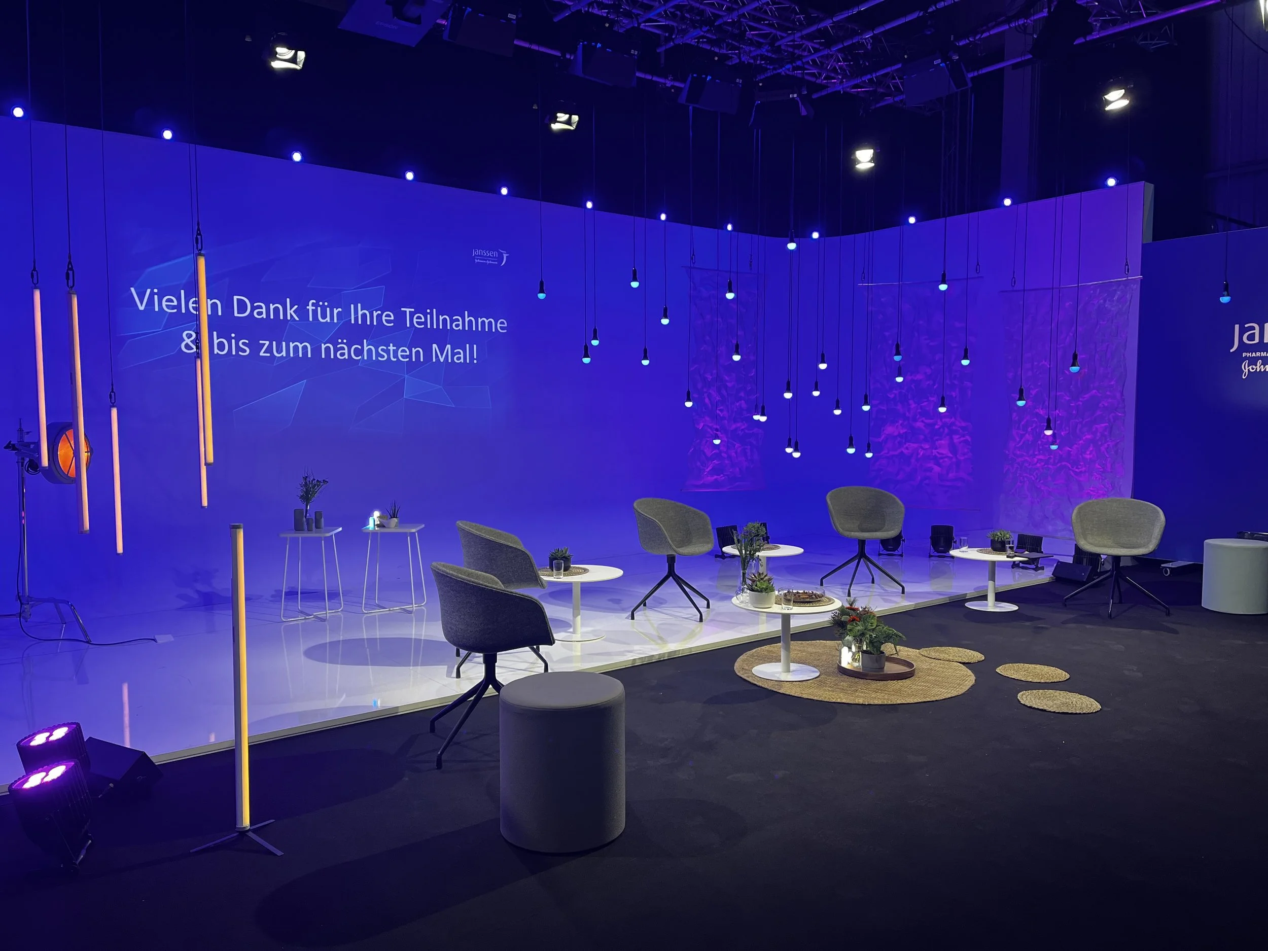 A staged setup with five gray chairs and four small white tables with potted plants on them. The backdrop is blue with hanging lights and a message in German.