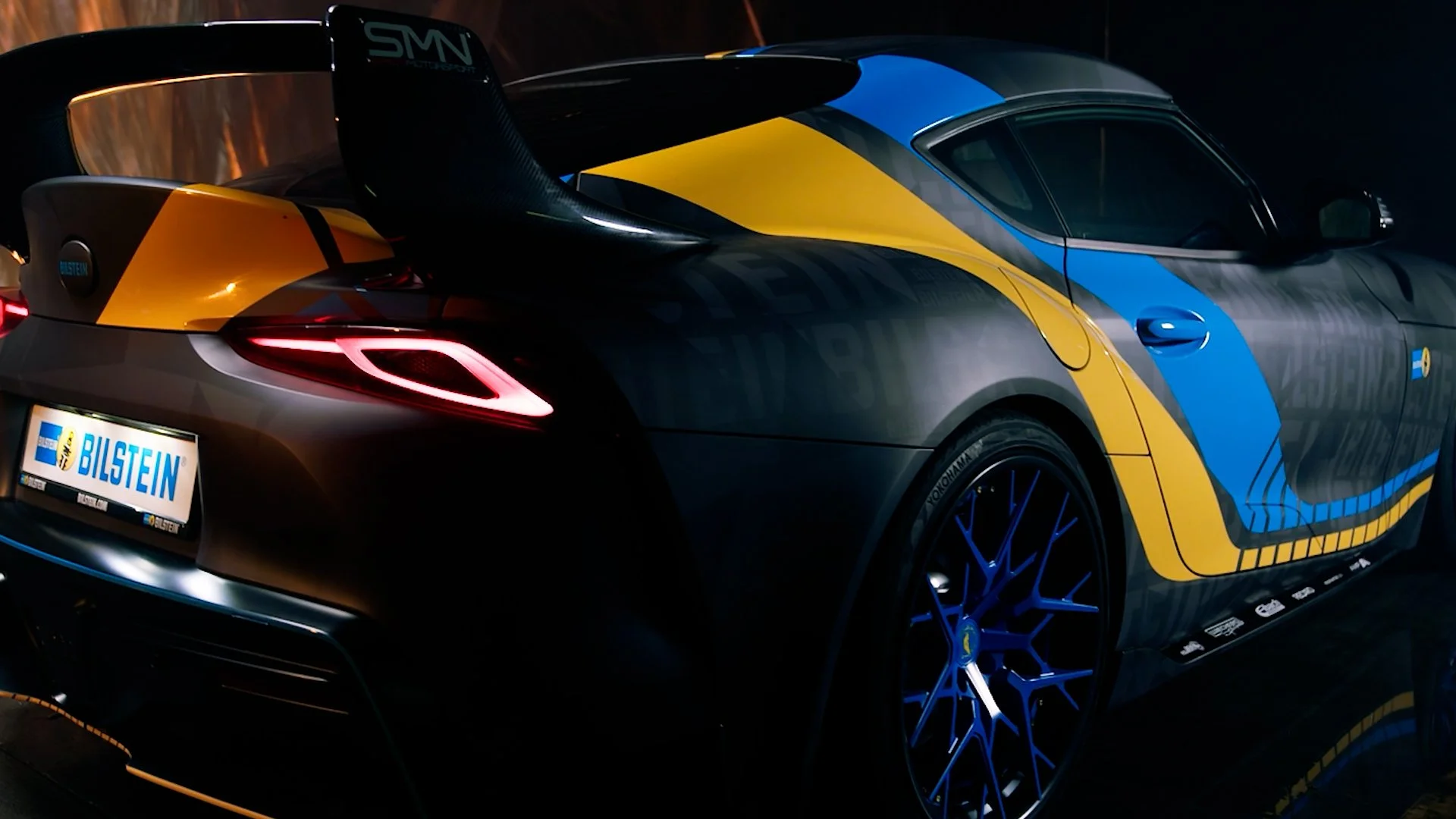 Close-up of a race car with a black, blue, and yellow design, rear lights illuminated, and a Bilstein license plate.