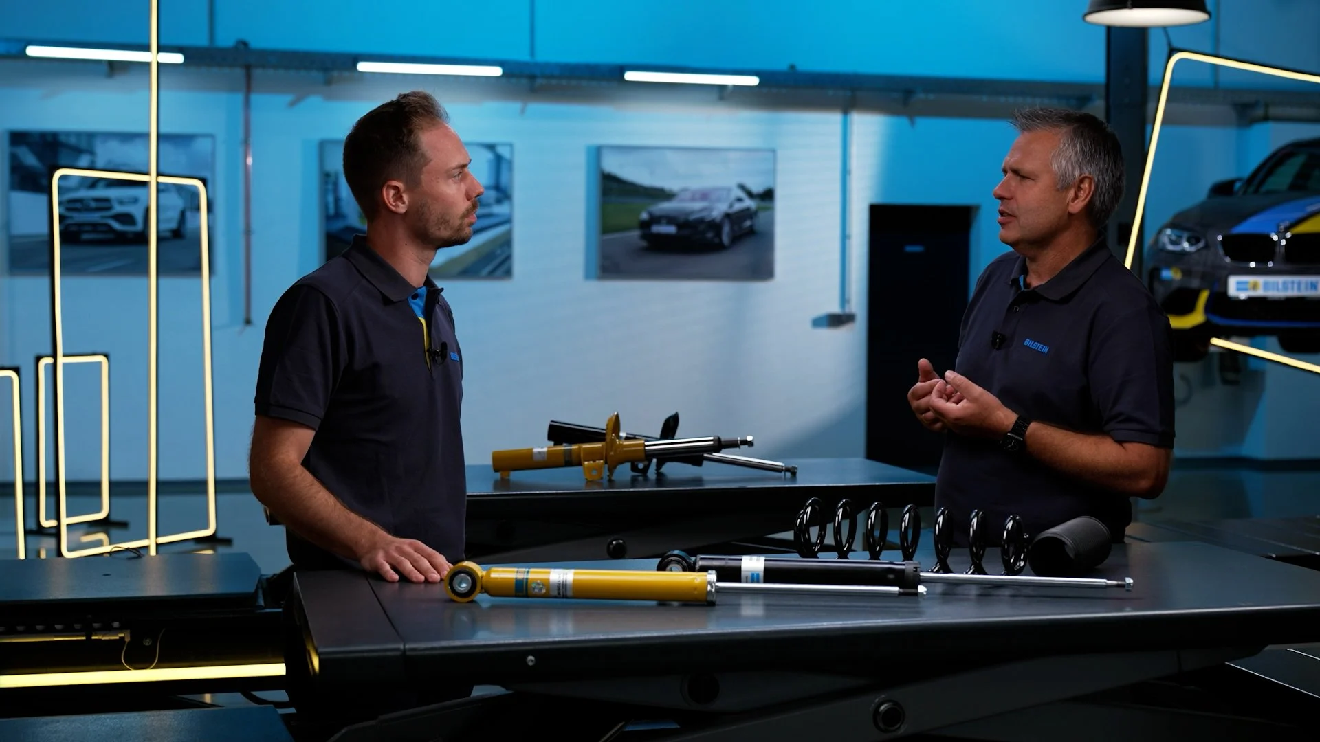 Two men in a discussion in a modern automotive workshop, with car suspension components on the table, including shock absorbers and springs, and cars and photos of cars on the walls in the background.