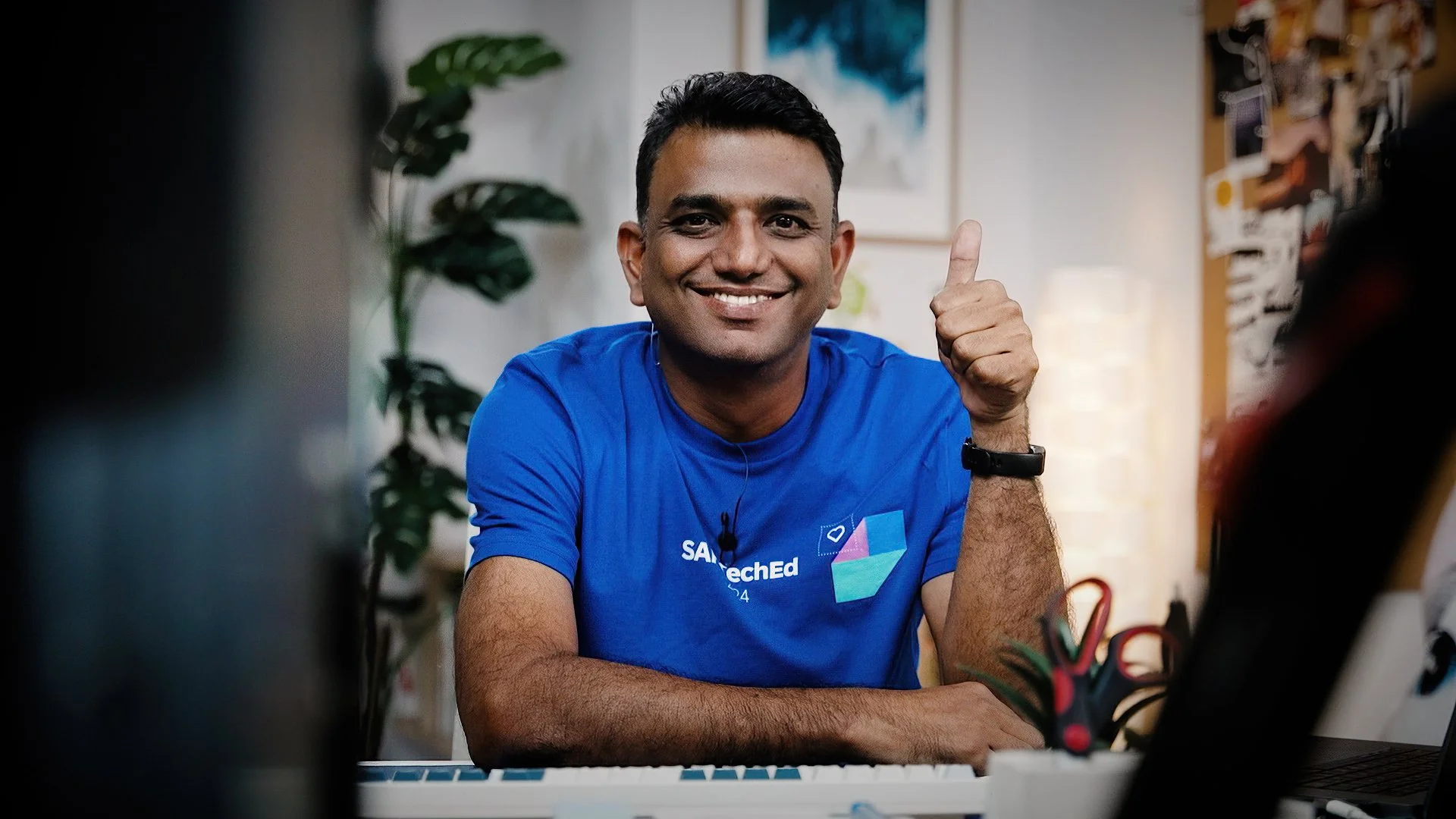 Smiling man in blue T-shirt giving a thumbs-up in a well-lit office or studio.