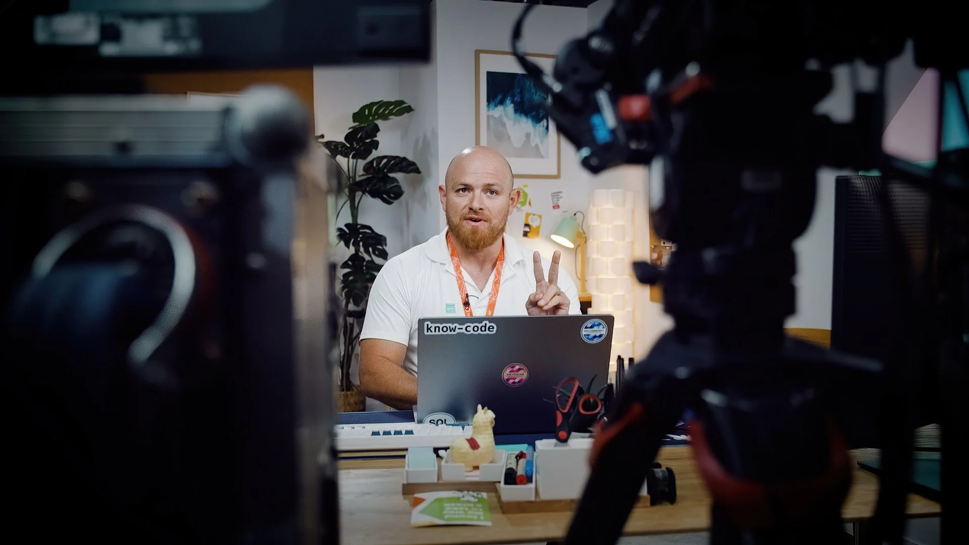 A man with a bald head and beard making a peace sign in front of a camera during a video recording or live stream in an office setting, with a laptop, desk stationery, and a framed picture on the wall in the background.