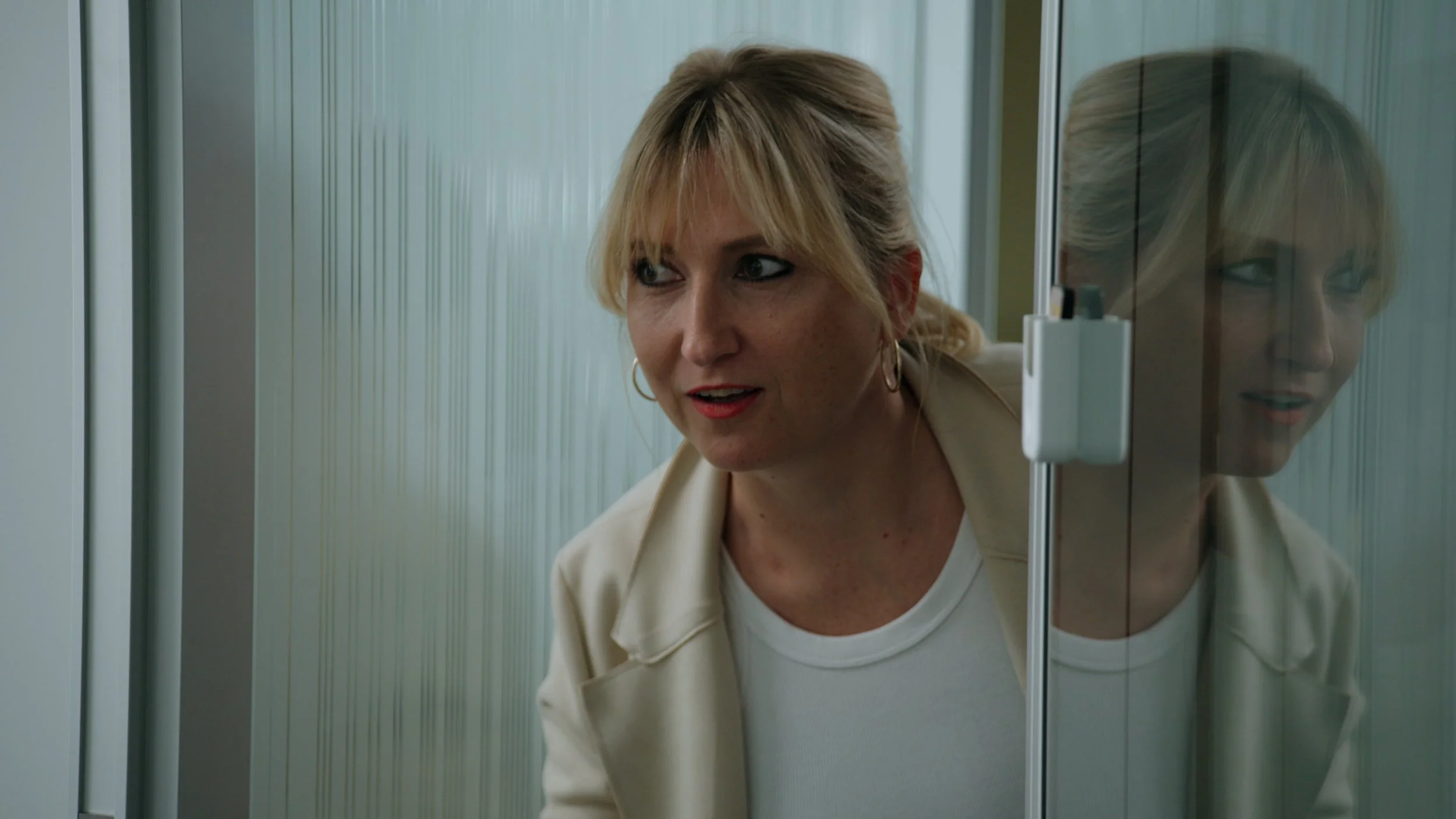 A woman with blonde hair in a ponytail peeking through an open door, with her reflection visible in a glass panel.