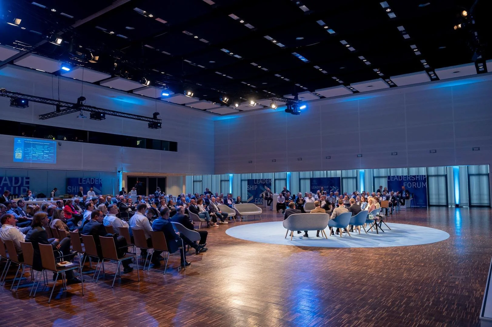 Large conference room filled with people attending a leadership forum, with a stage and seating area in the center, blue lighting, and a modern design.