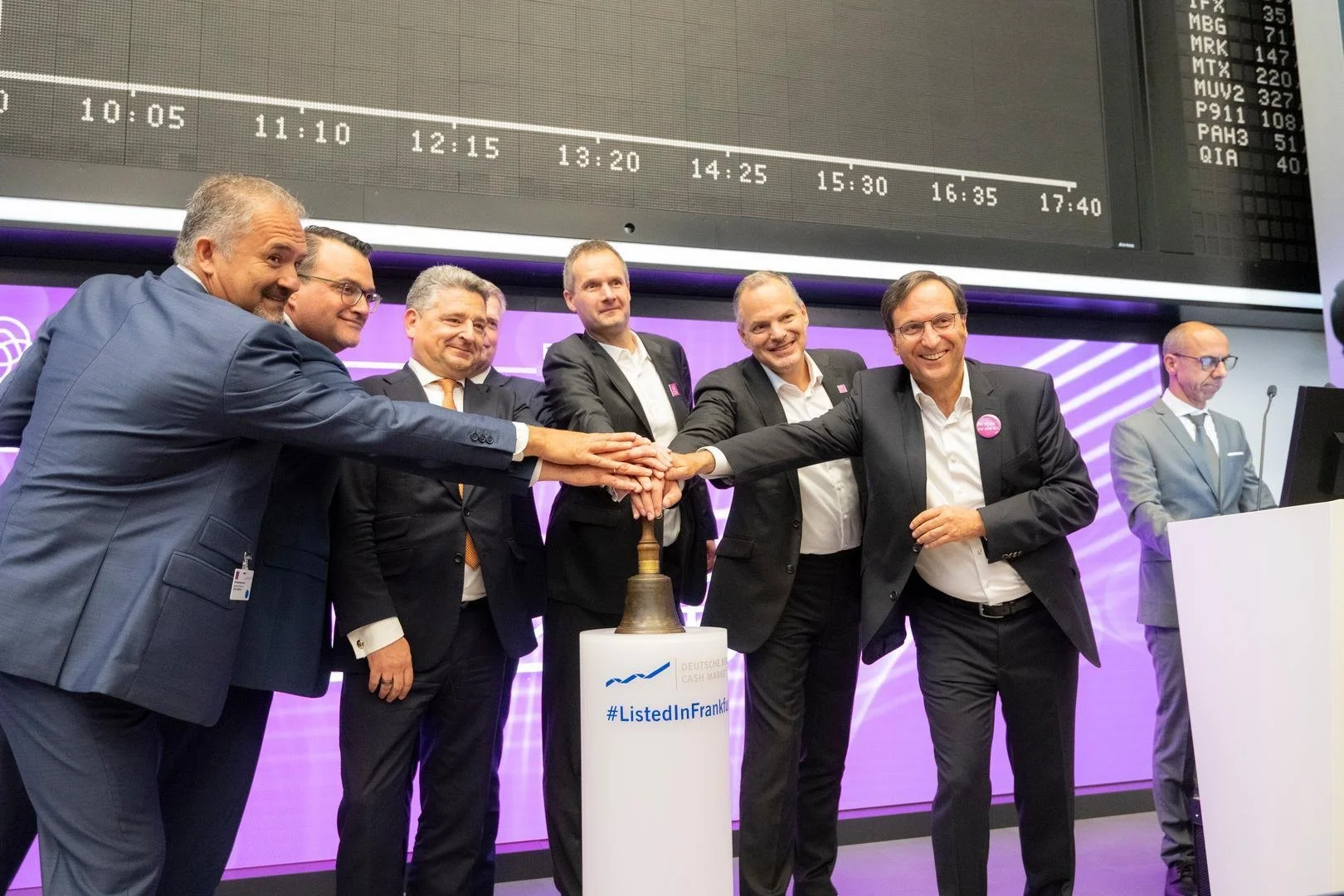 Group of seven men in business suits participating in a handshake gesture at a formal event, standing around a bell on a stand with a hashtag #ListedInFrankfurt, in front of a purple-lit backdrop and a large screen displaying a timetable.
