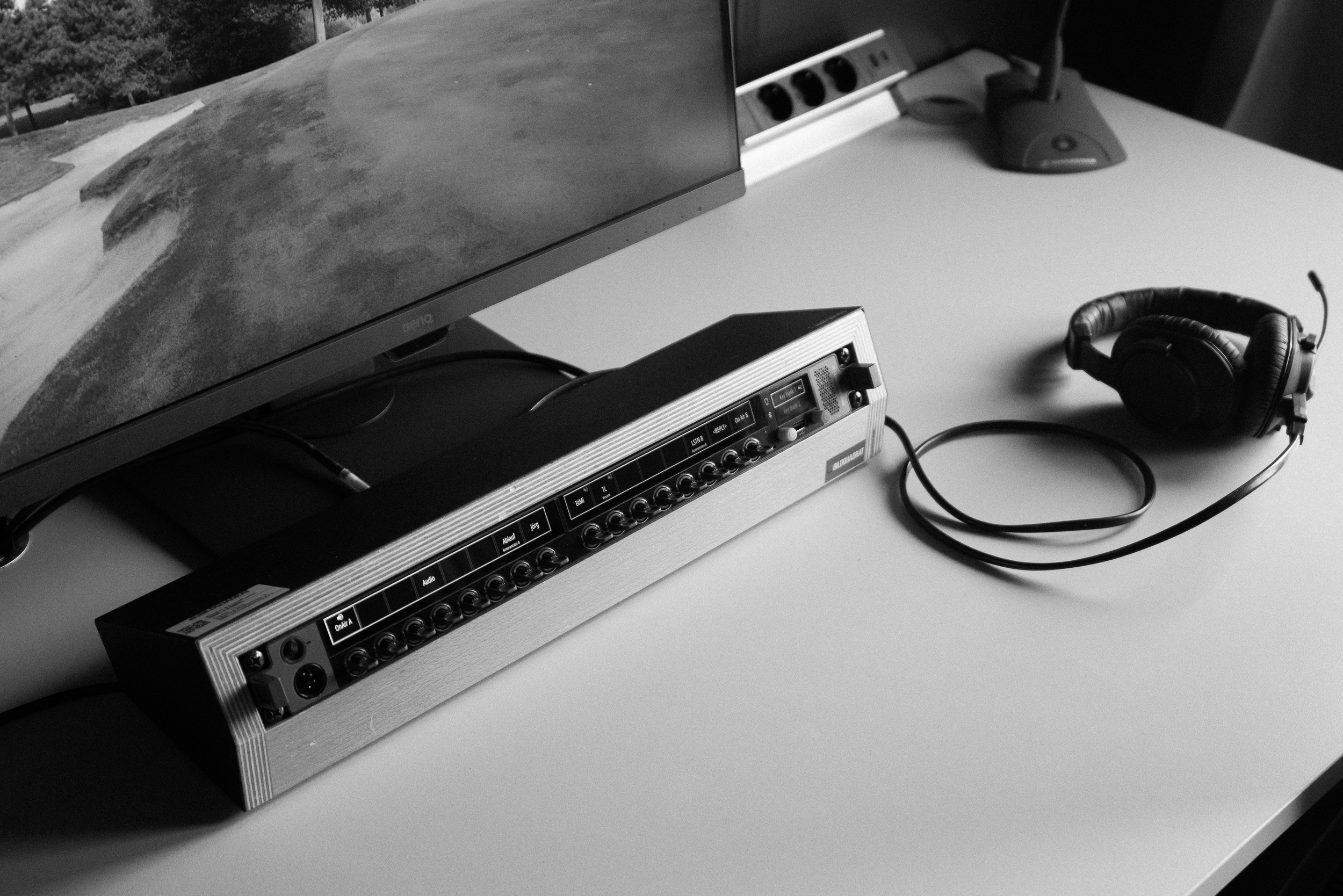 A computer monitor, a gray audio mixer with multiple input ports, a pair of black over-ear headphones, and a white power strip with black sockets on a white desk.