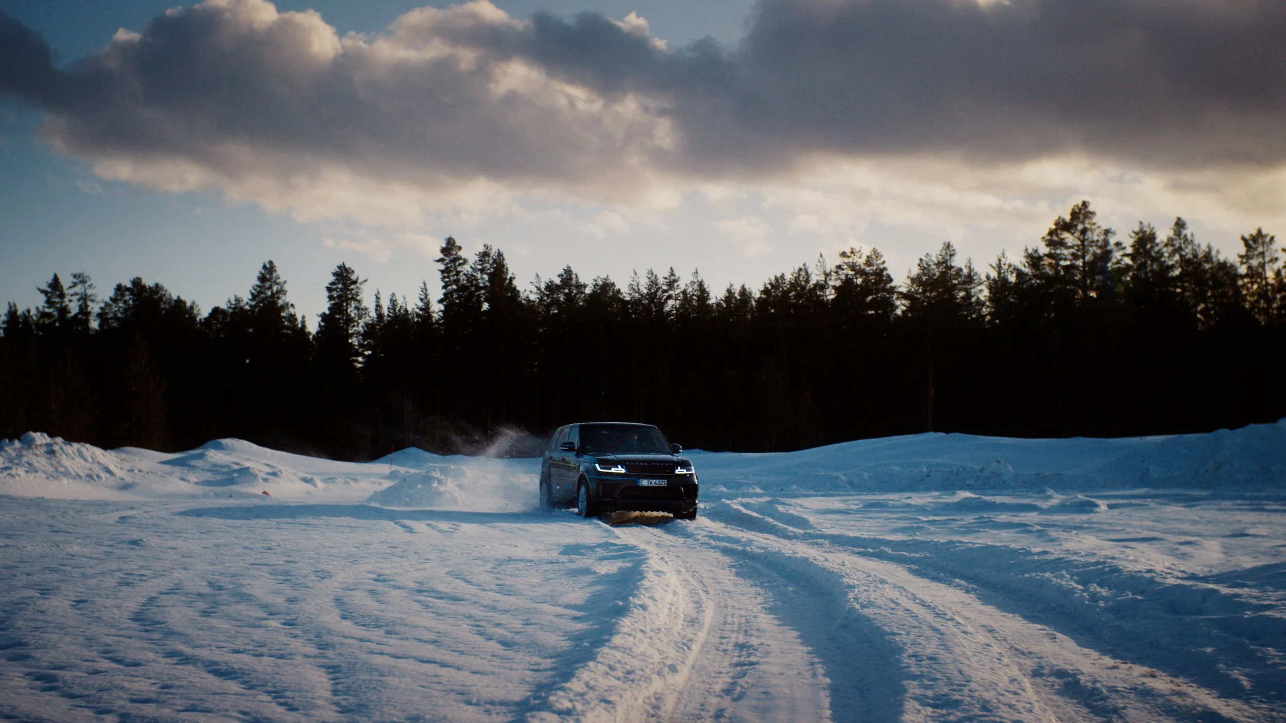 A black SUV driving on a snowy landscape with tire tracks, winter trees in the background, and a cloudy sky overhead.