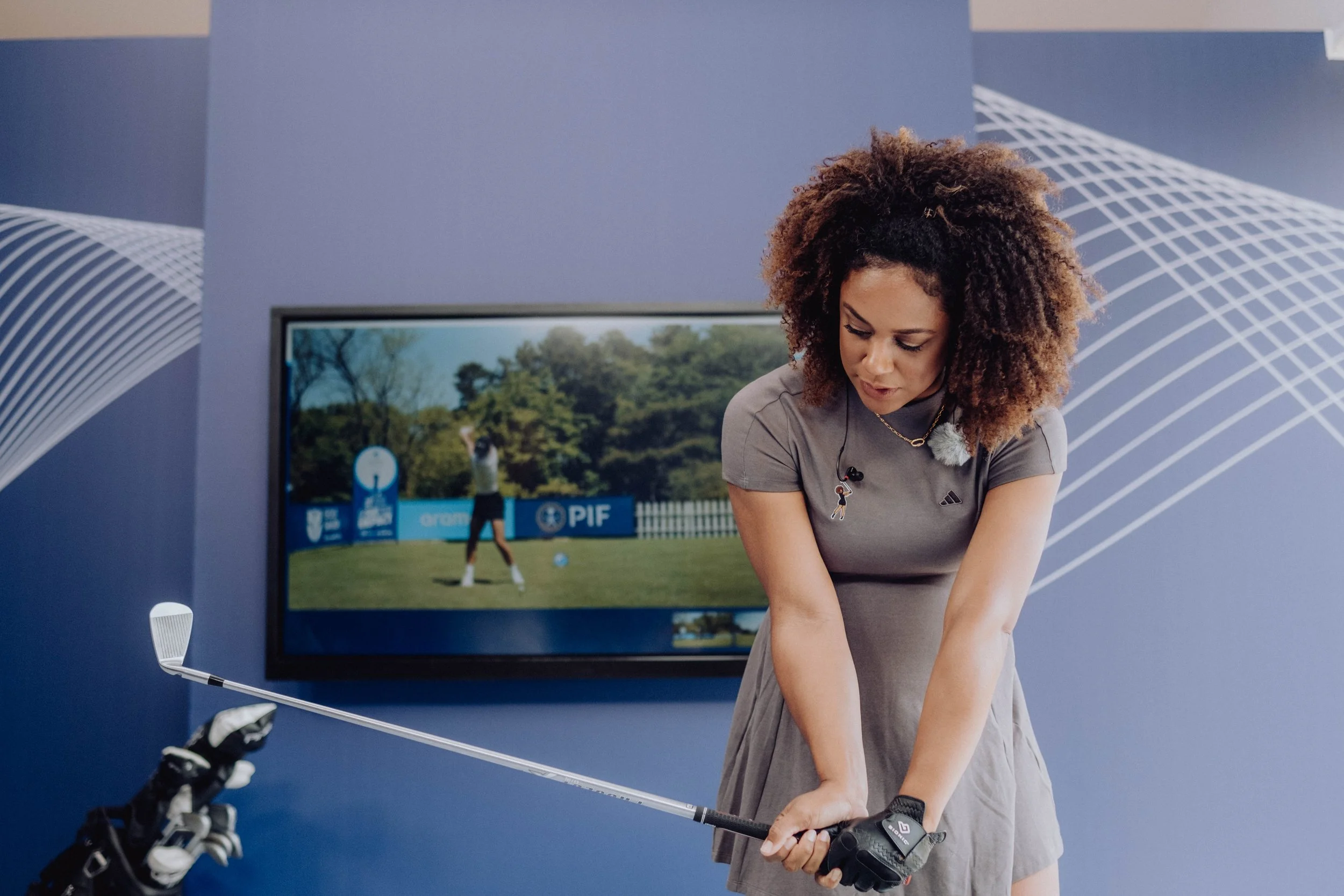 Woman practicing golf indoors with a golf club, TV screen in the background showing a golfer on a course.