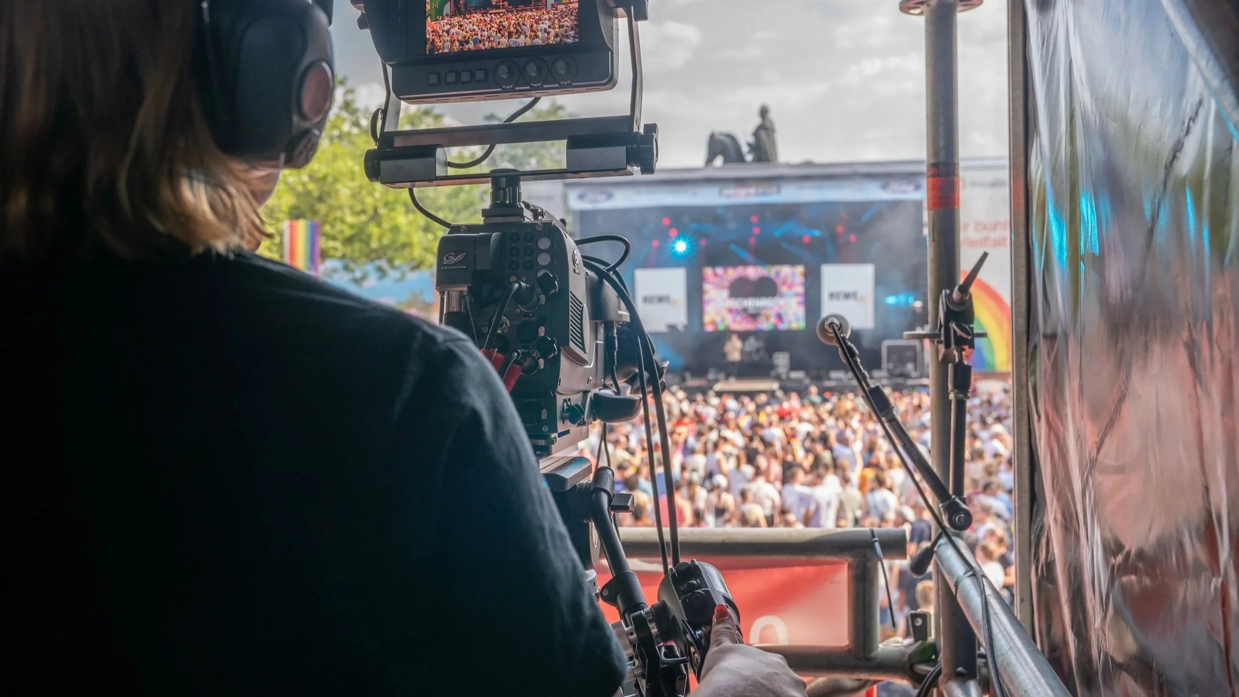 A camera operator is filming a large outdoor concert stage filled with a crowd under a cloudy sky.