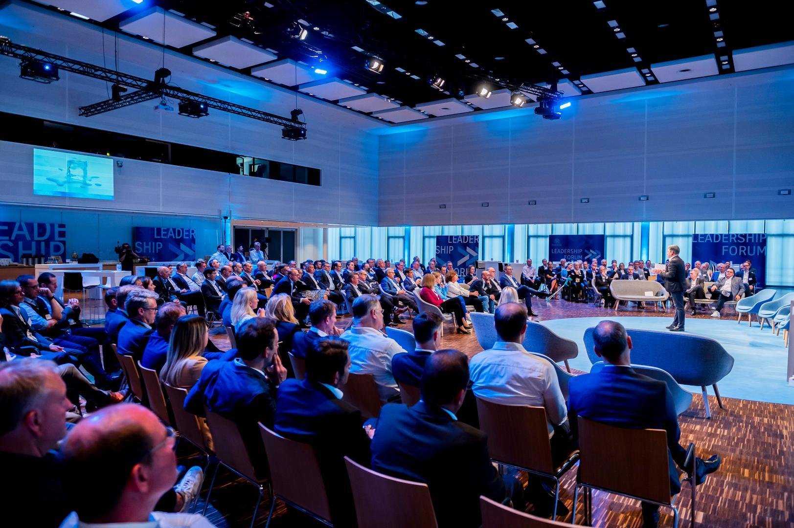 A conference room filled with seated attendees listening to a speaker on stage. The room has high ceilings, blue lighting, and banners reading 'Leadership Forum.'
